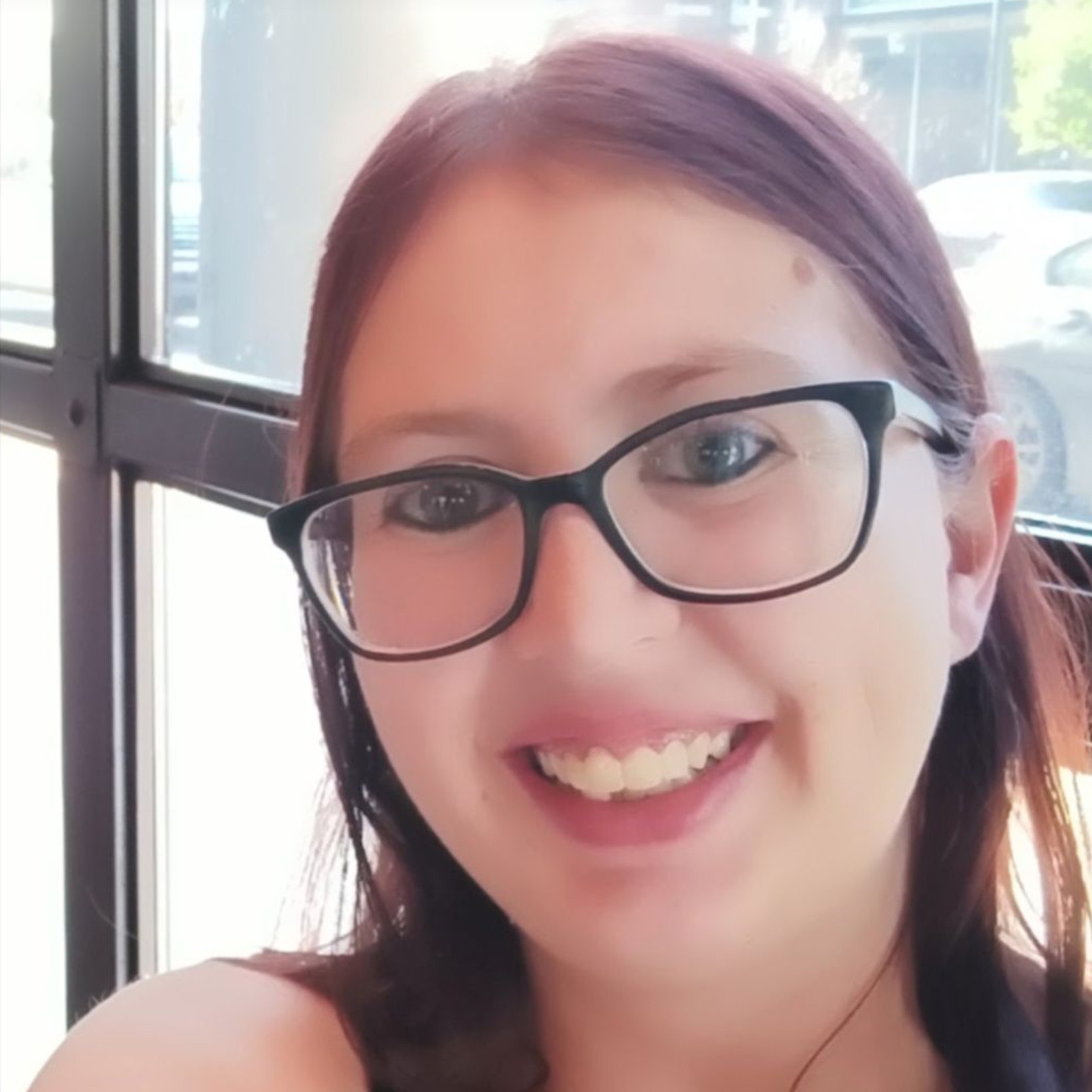 Woman with glasses smiles at the camera, inside a building near a window.