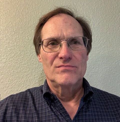 Man with glasses, looking forward, wearing a dark blue plaid shirt, against a light green wall.