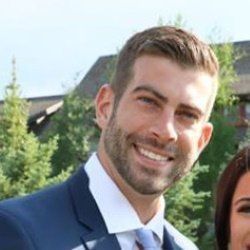 Man in blue suit, smiling outdoors, trees and building in the background.