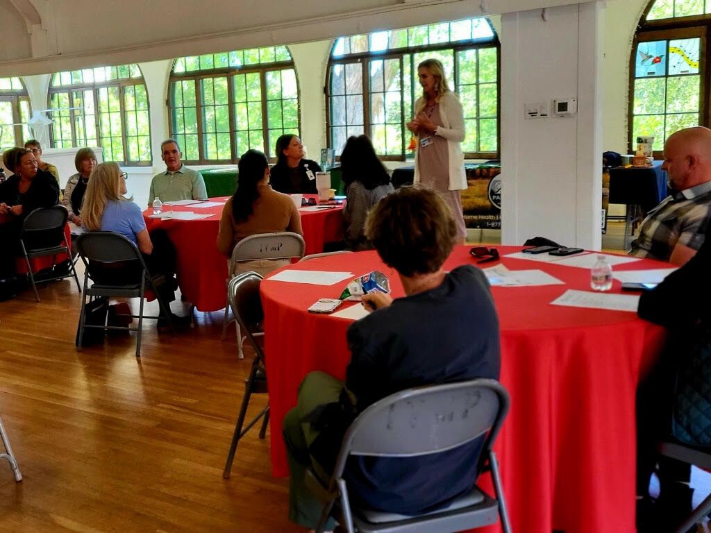 A woman speaks to a small group seated at red tables. The room has arched windows and wood floors.