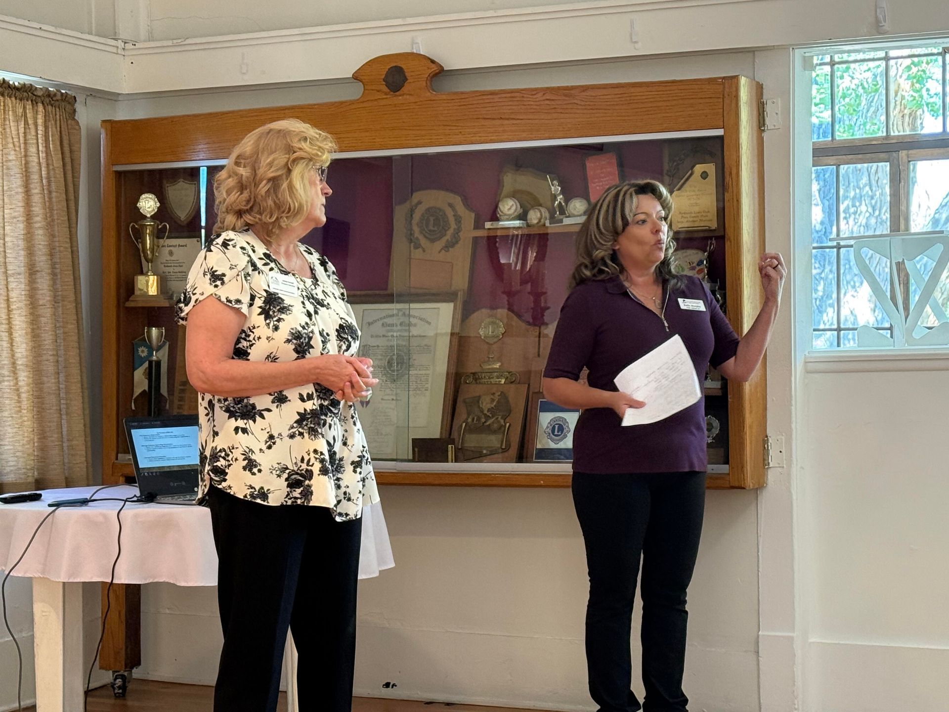 Two women stand near a display case in a room, one speaking, the other listening.