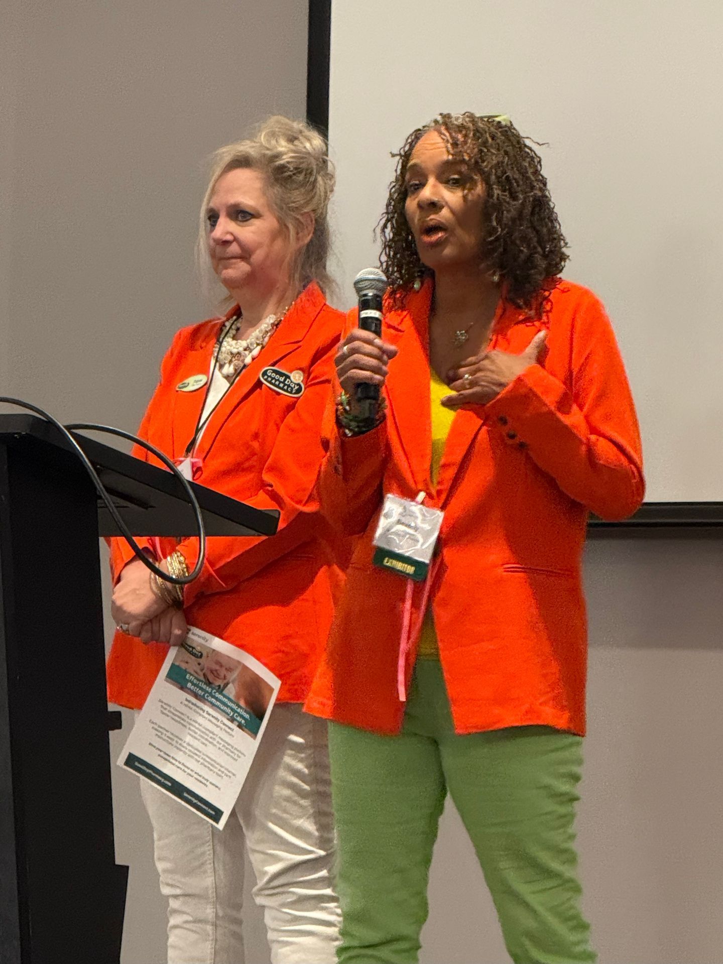Two women in orange blazers on a stage. One speaks into a microphone, the other holds a paper.
