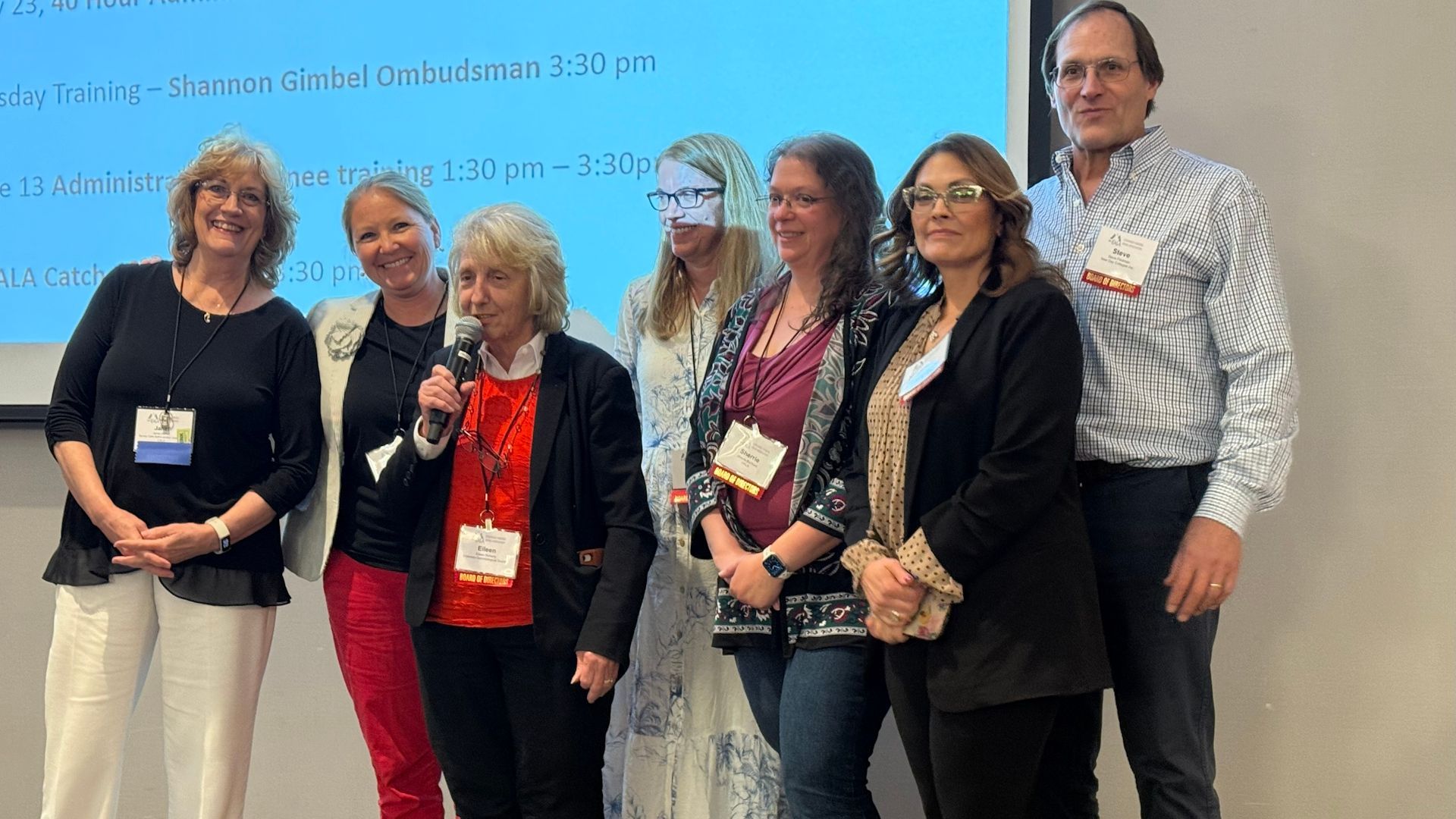 Group of people standing in front of a screen, one holding a microphone.