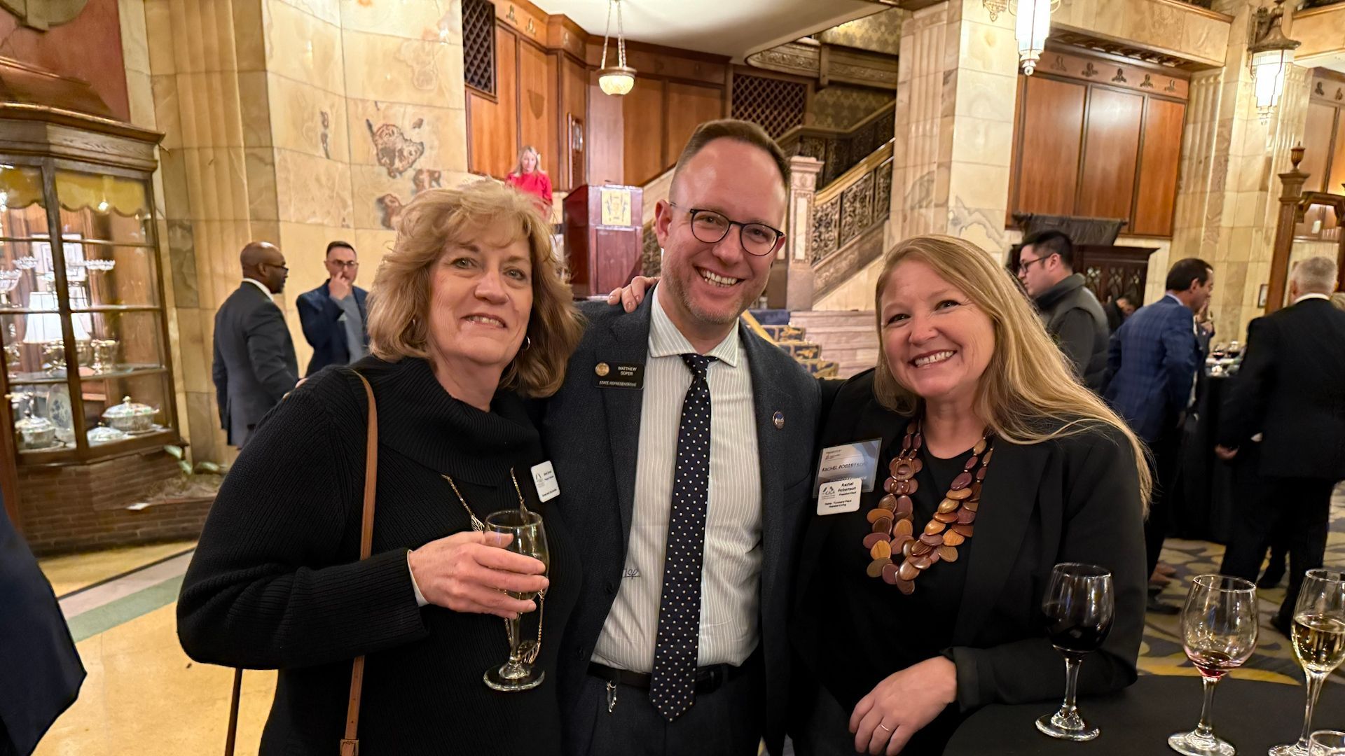 Three people smiling, posing together at an event. A man in a suit, two women with drinks. Interior with ornate architecture.