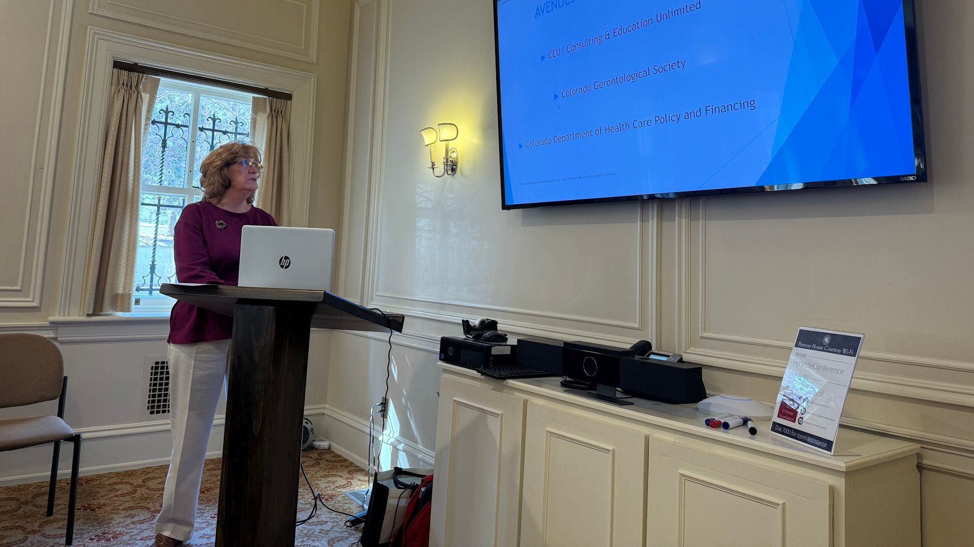Woman presenting at a podium, looking at a laptop, with a screen displaying text. Inside a room.