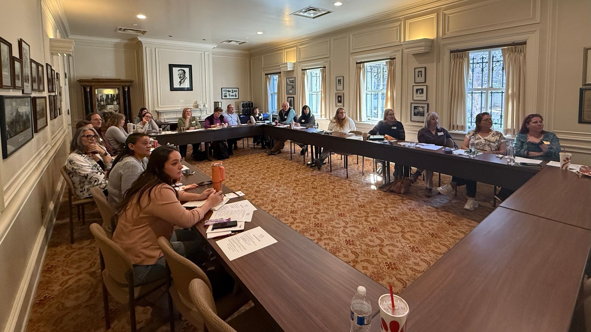 Group of people sitting at a long table in a formal room, possibly a meeting.