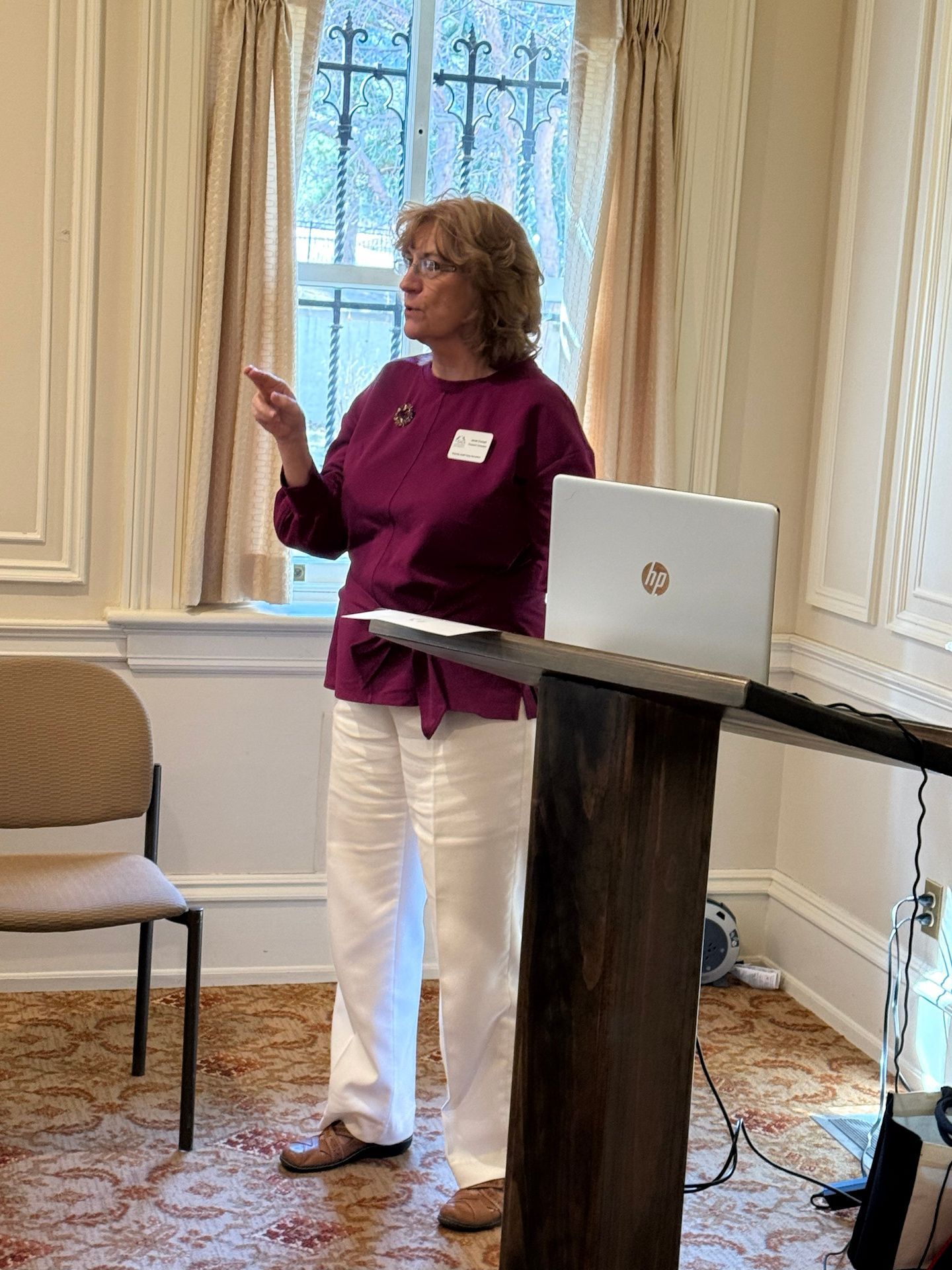Woman in burgundy top and white pants gestures at a presentation, standing near a laptop on a wooden podium.