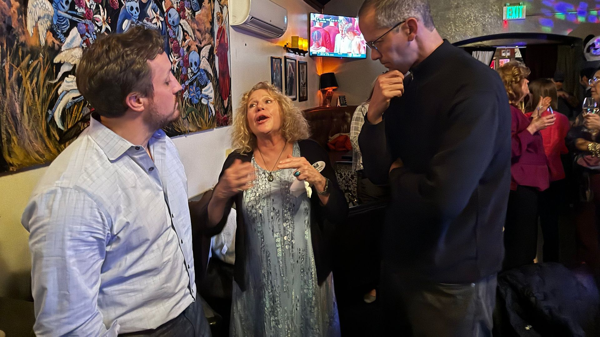 Three people talking in a bar, woman in sparkly dress gesturing, men in button-up shirt and dark sweater.