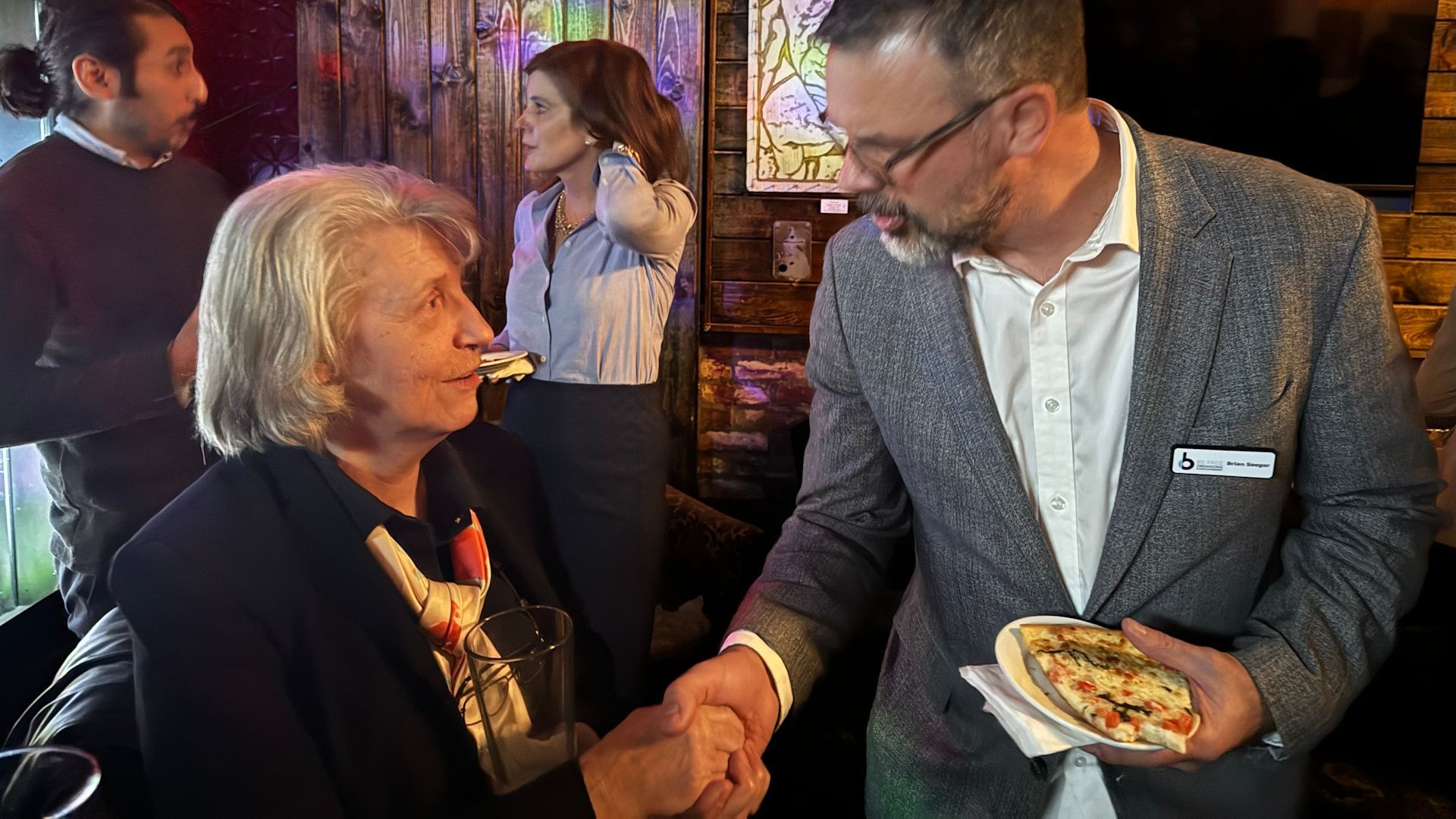 Man shaking hands with older woman, both smiling, at an event. Other guests are in background.