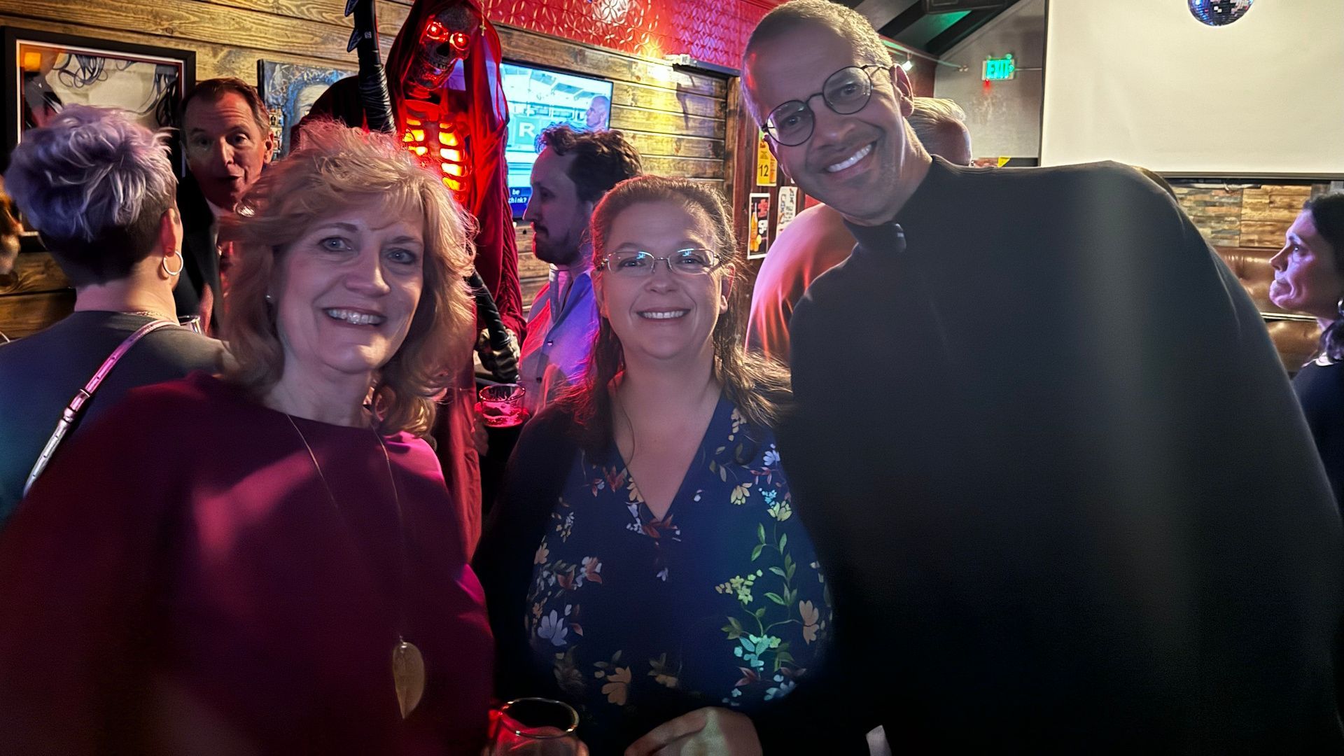 Three people smiling and posing for a photo in a dimly lit bar, surrounded by other patrons.