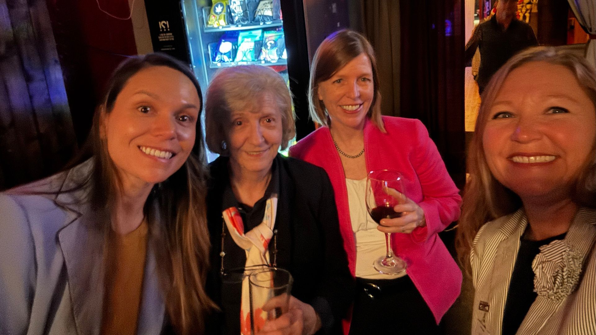 Four smiling people taking a selfie inside, one holding a drink, with a vending machine in the background.