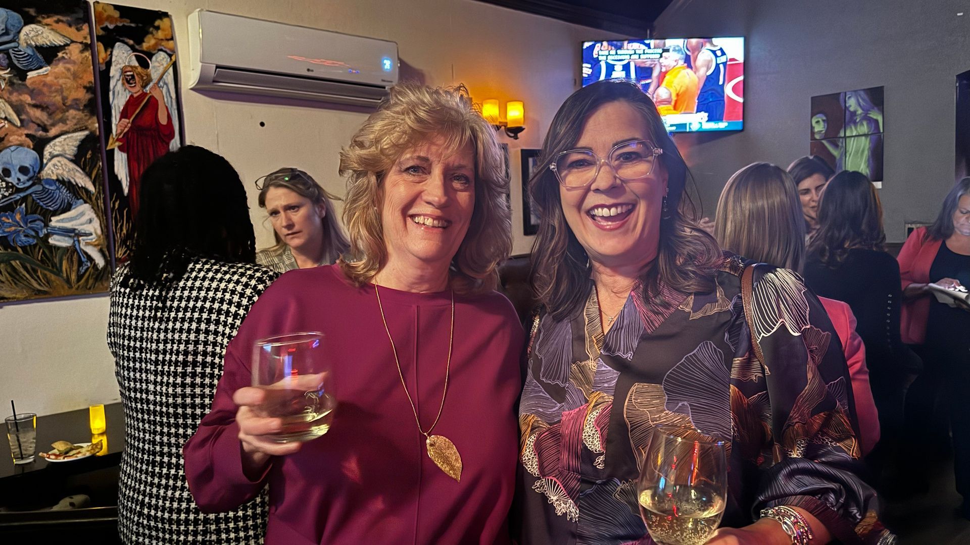 Two women smiling, holding drinks, indoors at a gathering. One in purple, other in paisley scarf.