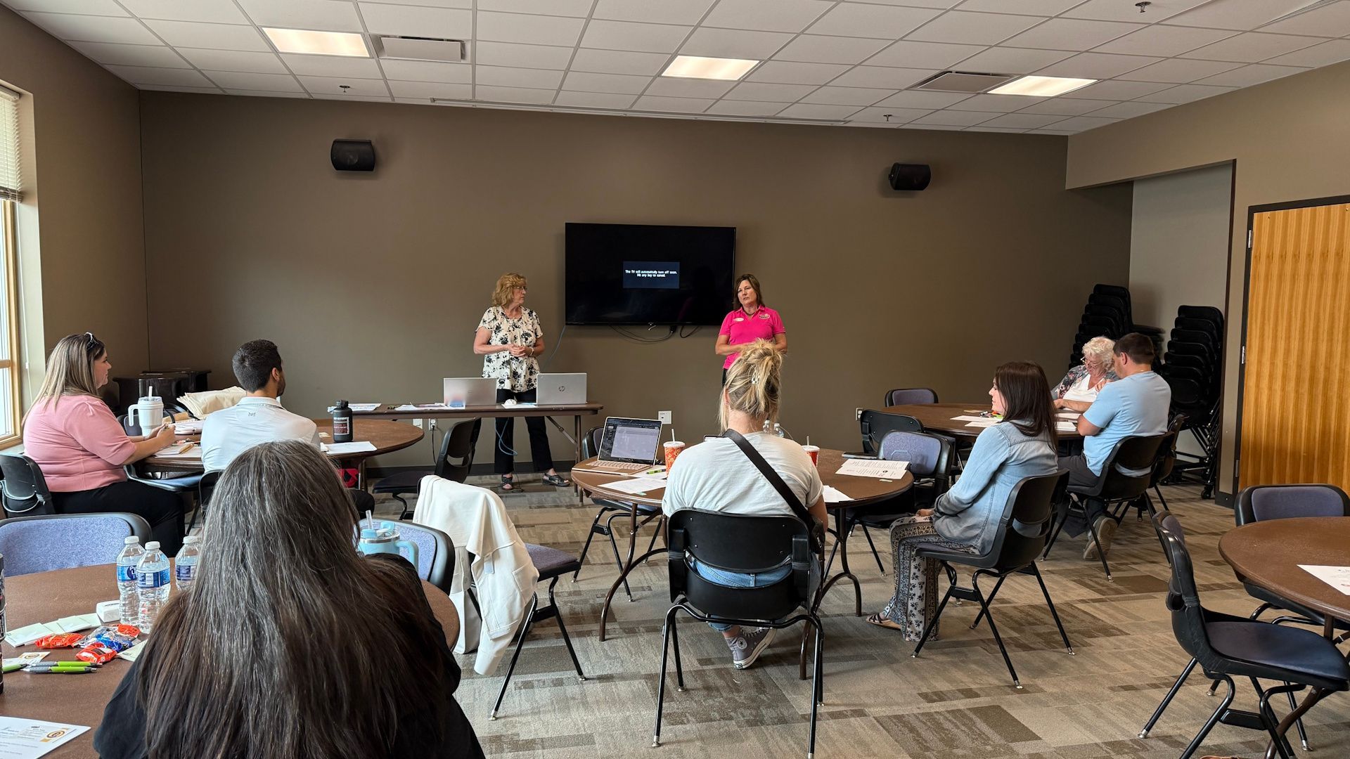 Two women presenting to a group seated at round tables in a conference room.