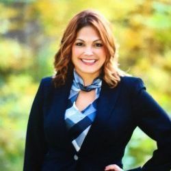 Woman in a blazer smiles, hand on hip; a blue and white patterned scarf, outdoor background.
