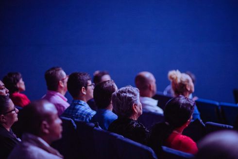 Audience members seated in a dark auditorium, gazing upwards toward the stage. Blue lighting.