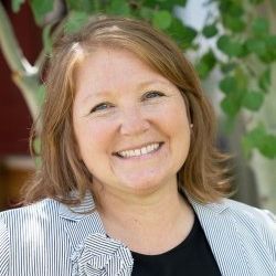 Woman with light brown hair, smiling, wearing a striped blazer, outdoors with trees.