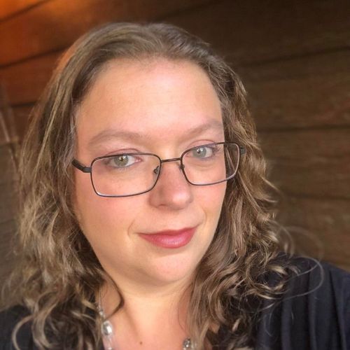 Woman with glasses and curly blonde hair smiles at the camera, wearing a dark shirt and necklace.
