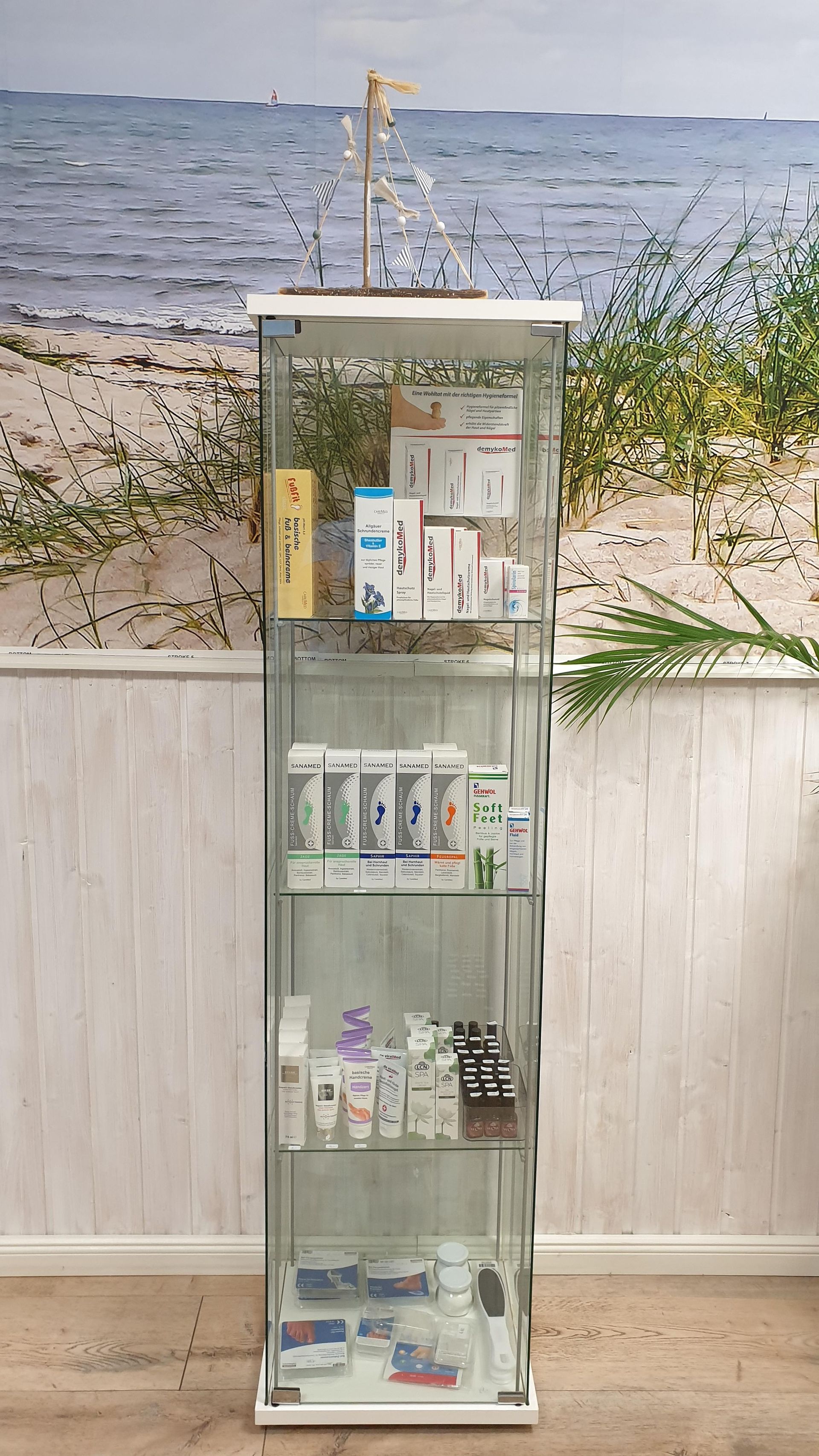 Eine Glasvitrine mit vielen Kosmetika und einem Bild vom Meer an der Wand.