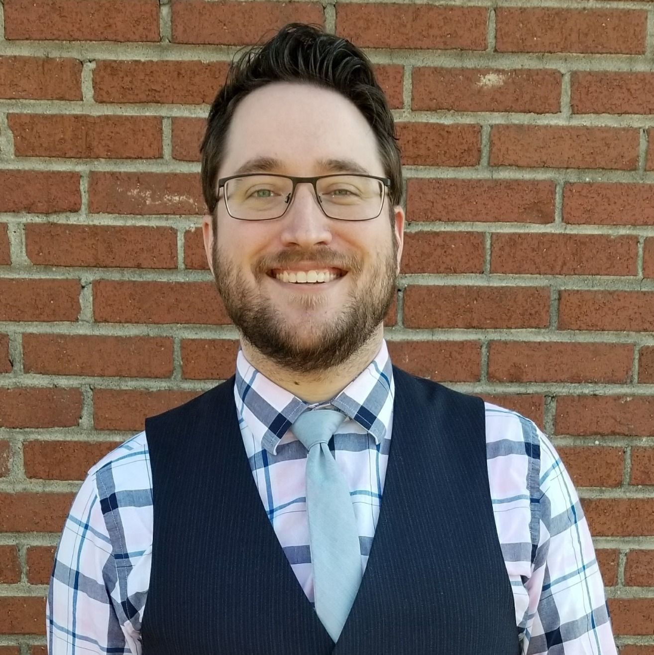 Man with glasses smiles in front of a brick wall, wearing a vest, tie and plaid shirt.