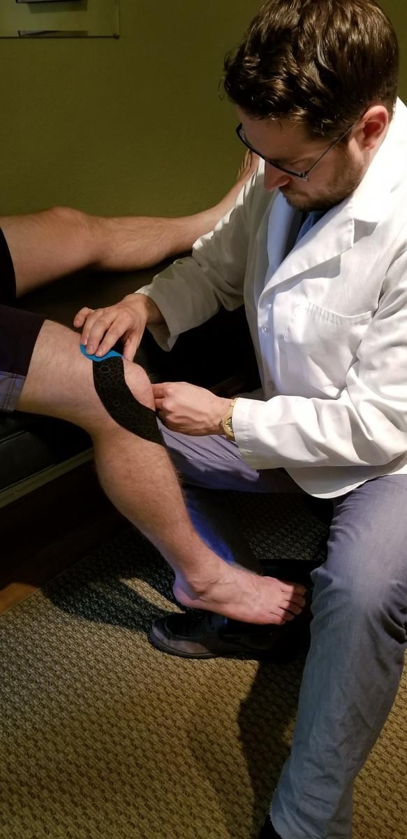 A person in a white coat applies kinesiology tape to another person's knee indoors.