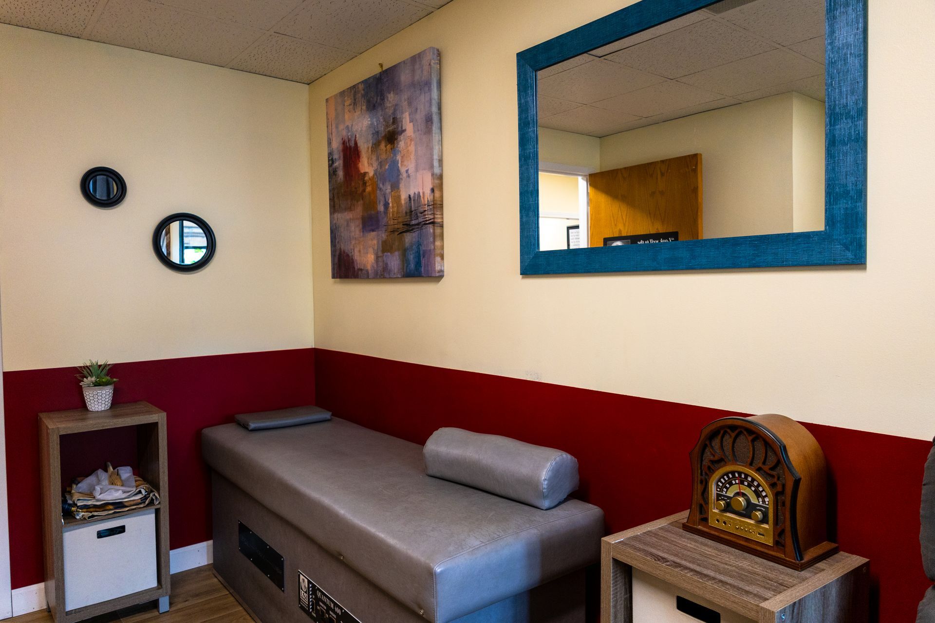A massage room with red and cream walls, a gray bed, mirror, and decorative art.