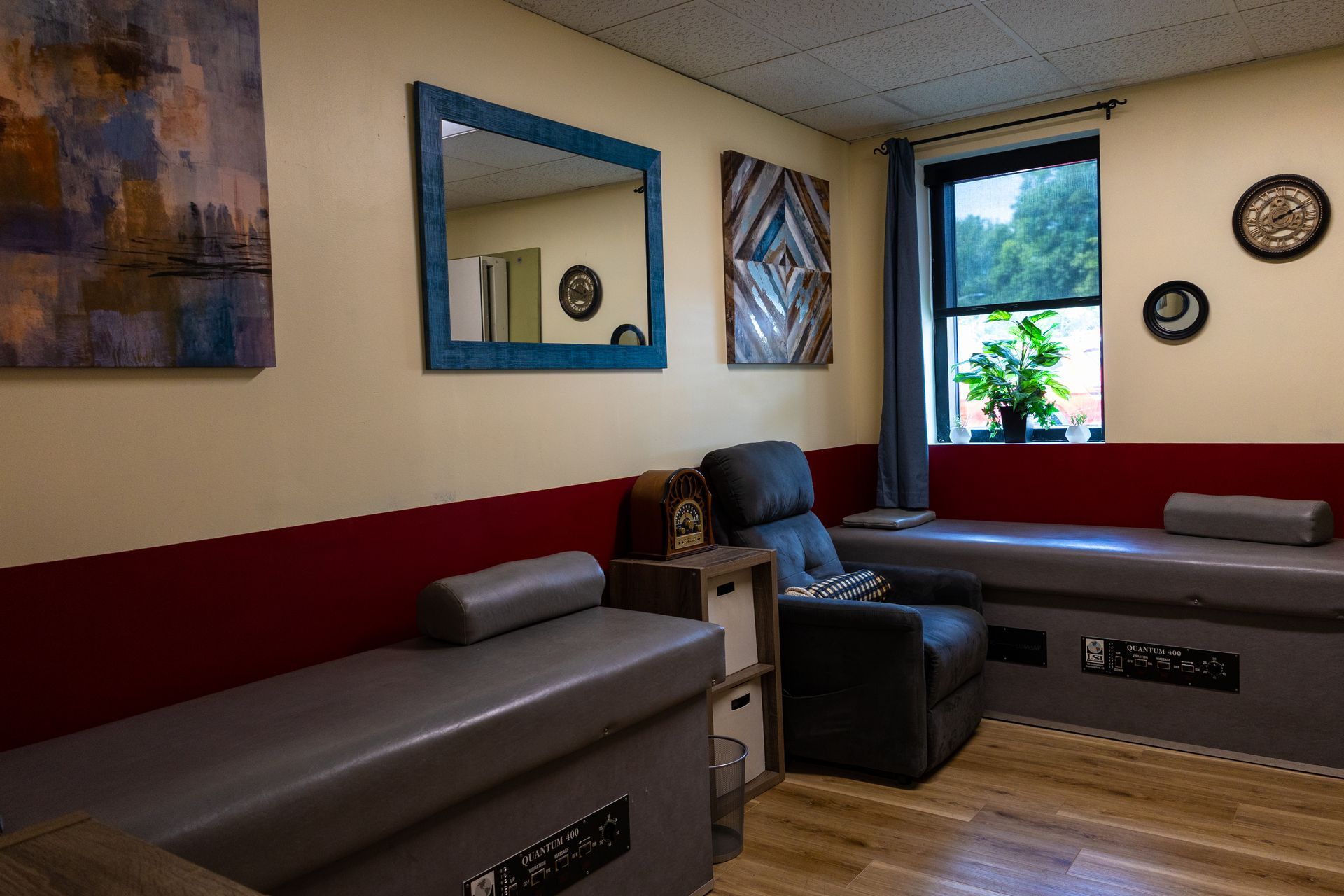 Therapy room with a recliner, benches, and a window with plants. Artwork and a mirror adorn the walls.