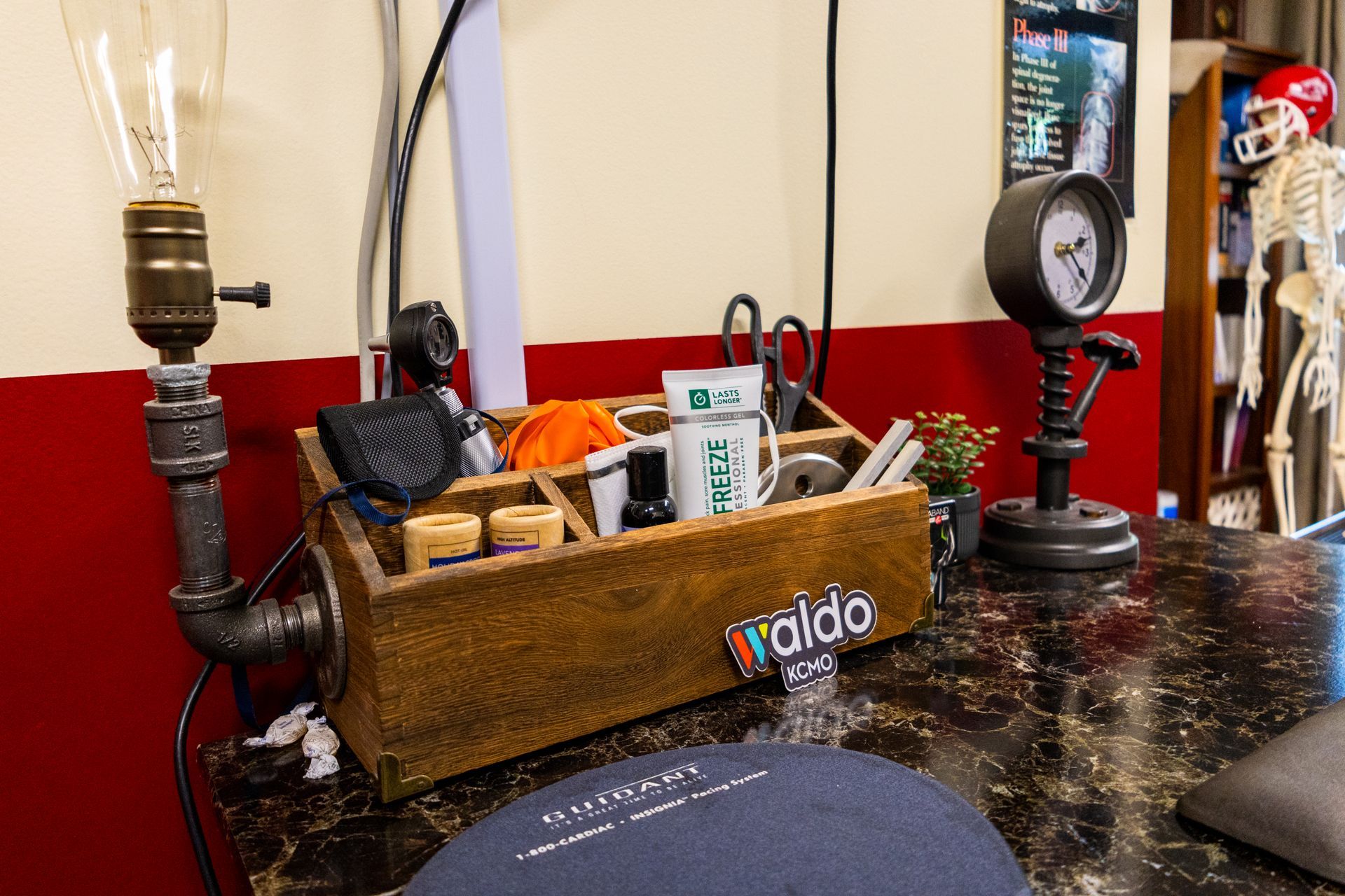 Desk with toolbox, lamp, gauge, and skeleton in a room with red and cream walls.