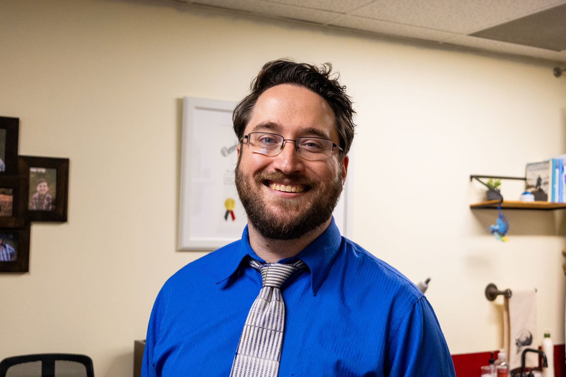 Man with glasses and beard smiles in a blue shirt, indoors, in front of a wall with framed art.