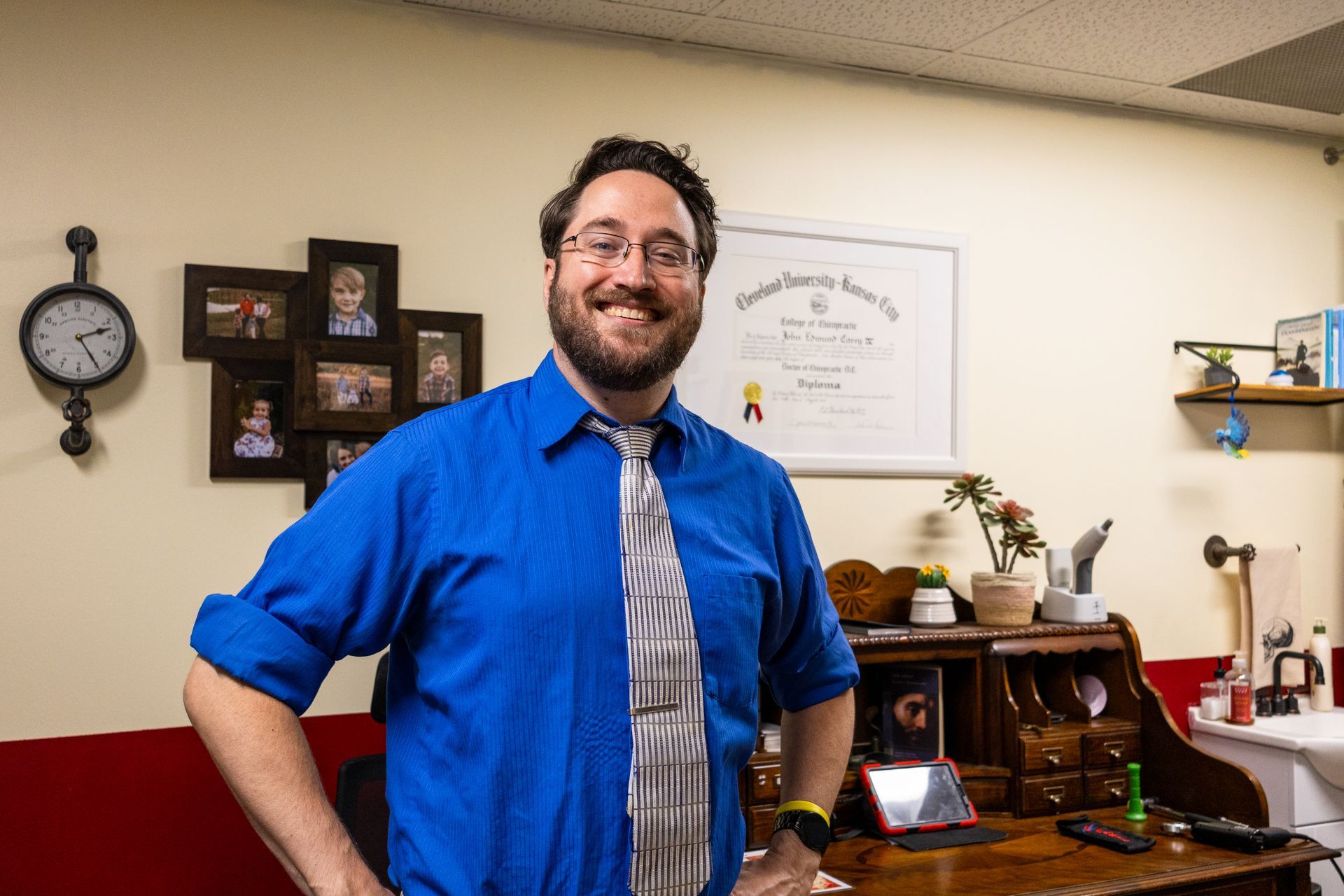 Man in blue shirt and tie smiles in front of a desk, diploma, and photos.