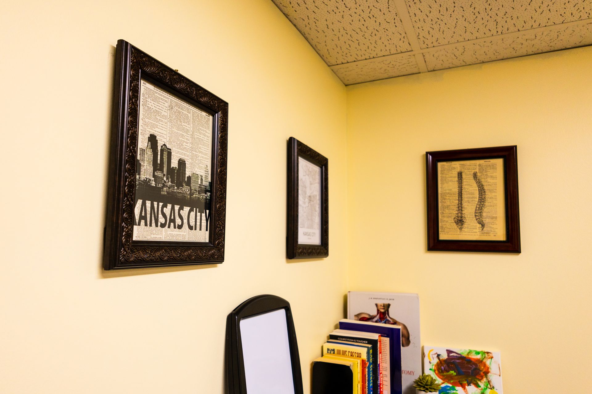 Gallery wall in a corner with three framed art pieces and a shelf of books, with a black mirror leaning on the shelf.