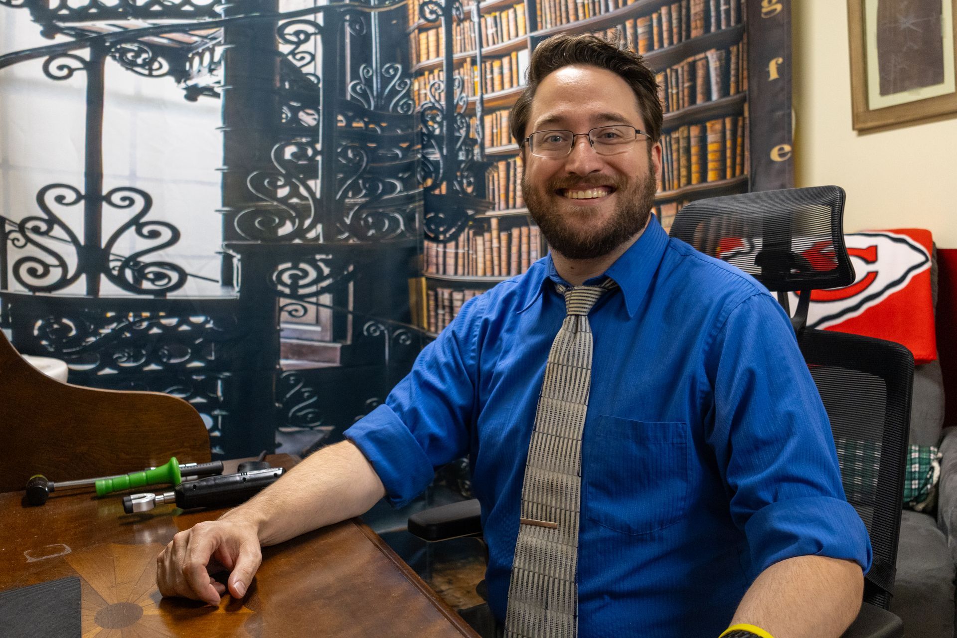 Man with glasses and beard smiles at desk in home office with spiral staircase decor.
