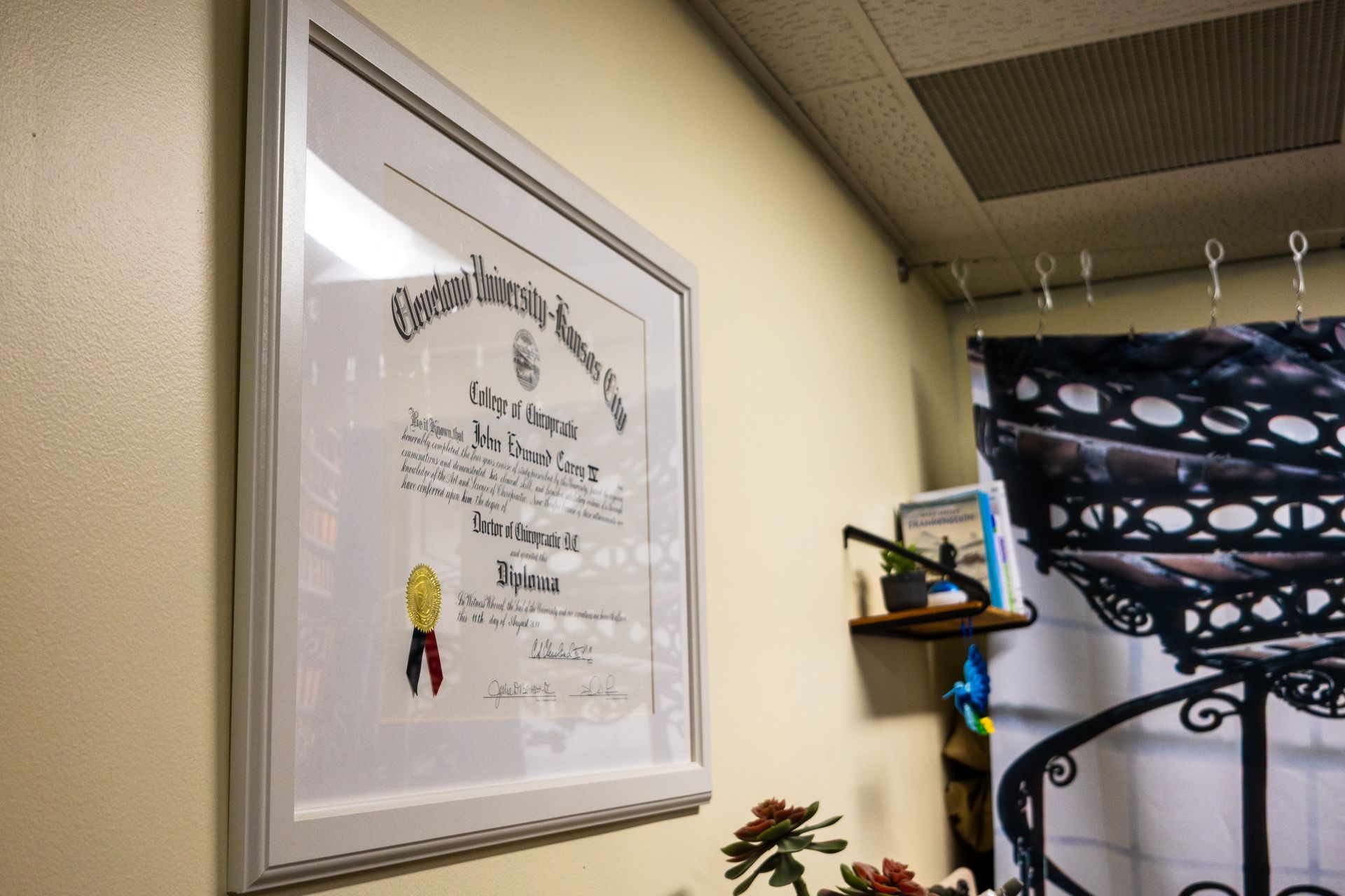 Framed diploma on a cream wall. A small shelf and decorative metalwork are also visible.