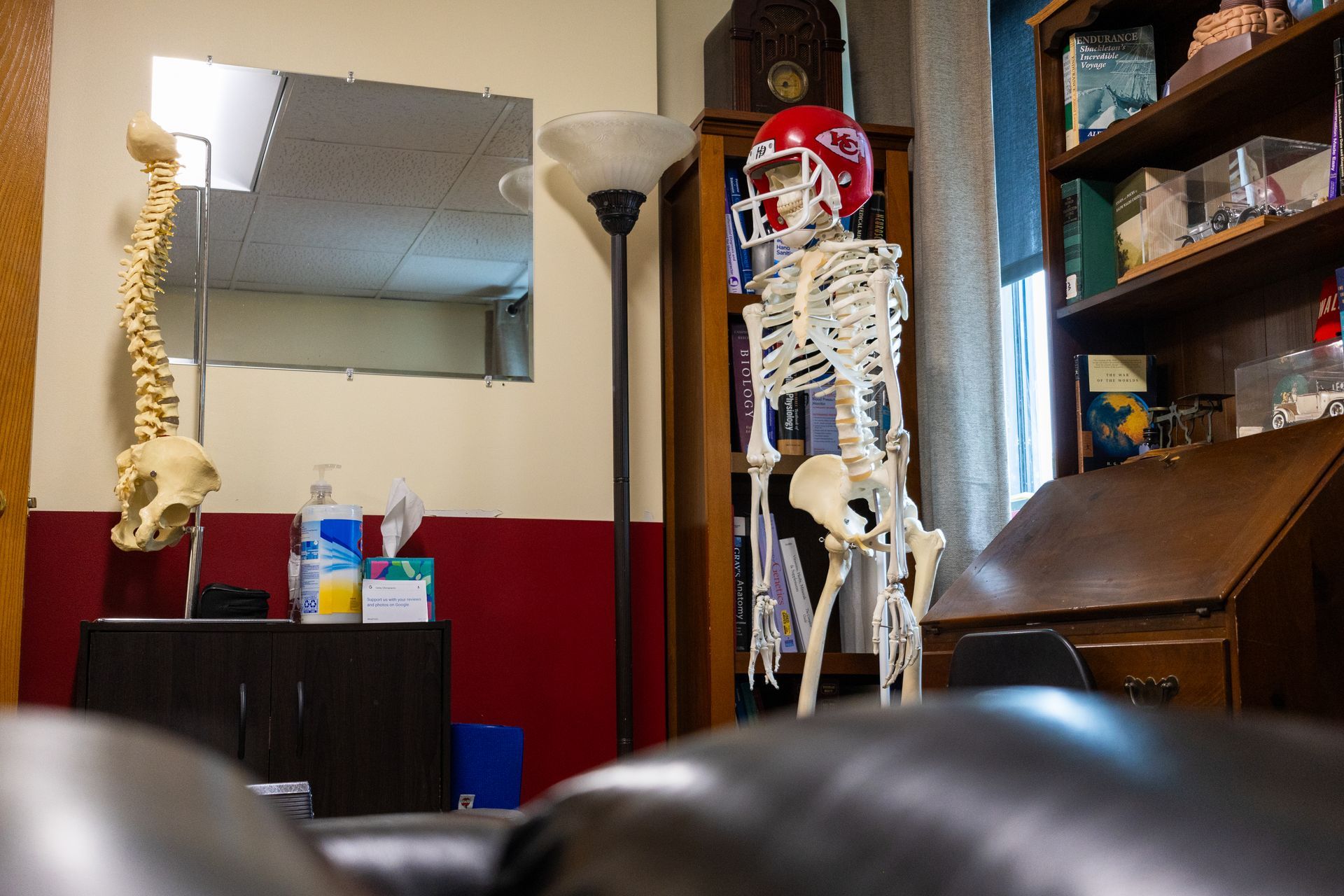 Skeleton wearing a red football helmet stands in an office, near a bookshelf and a hanging spine model.