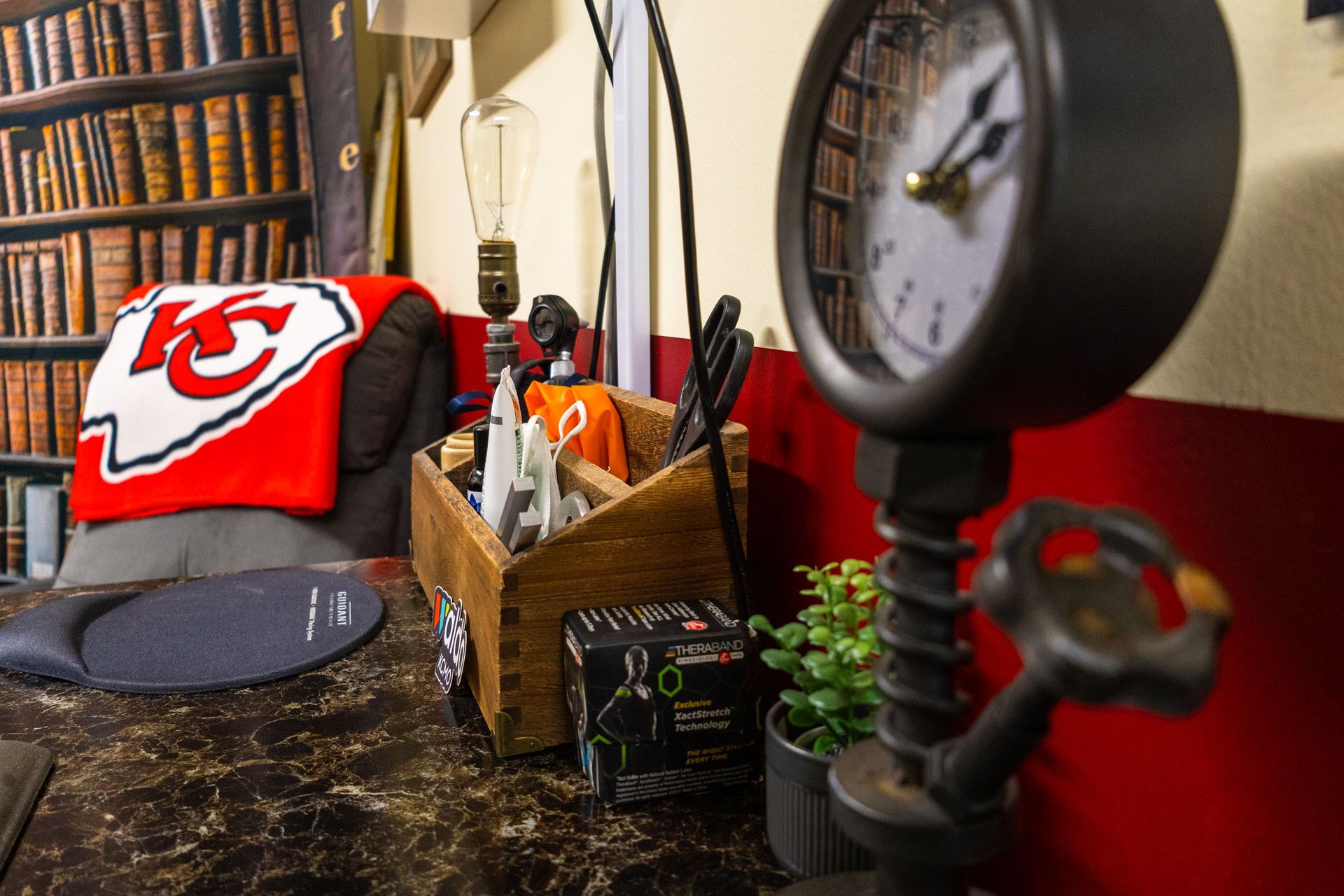 Desk with clock, pen holder, lamp, Chiefs blanket, and bookcase wallpaper.