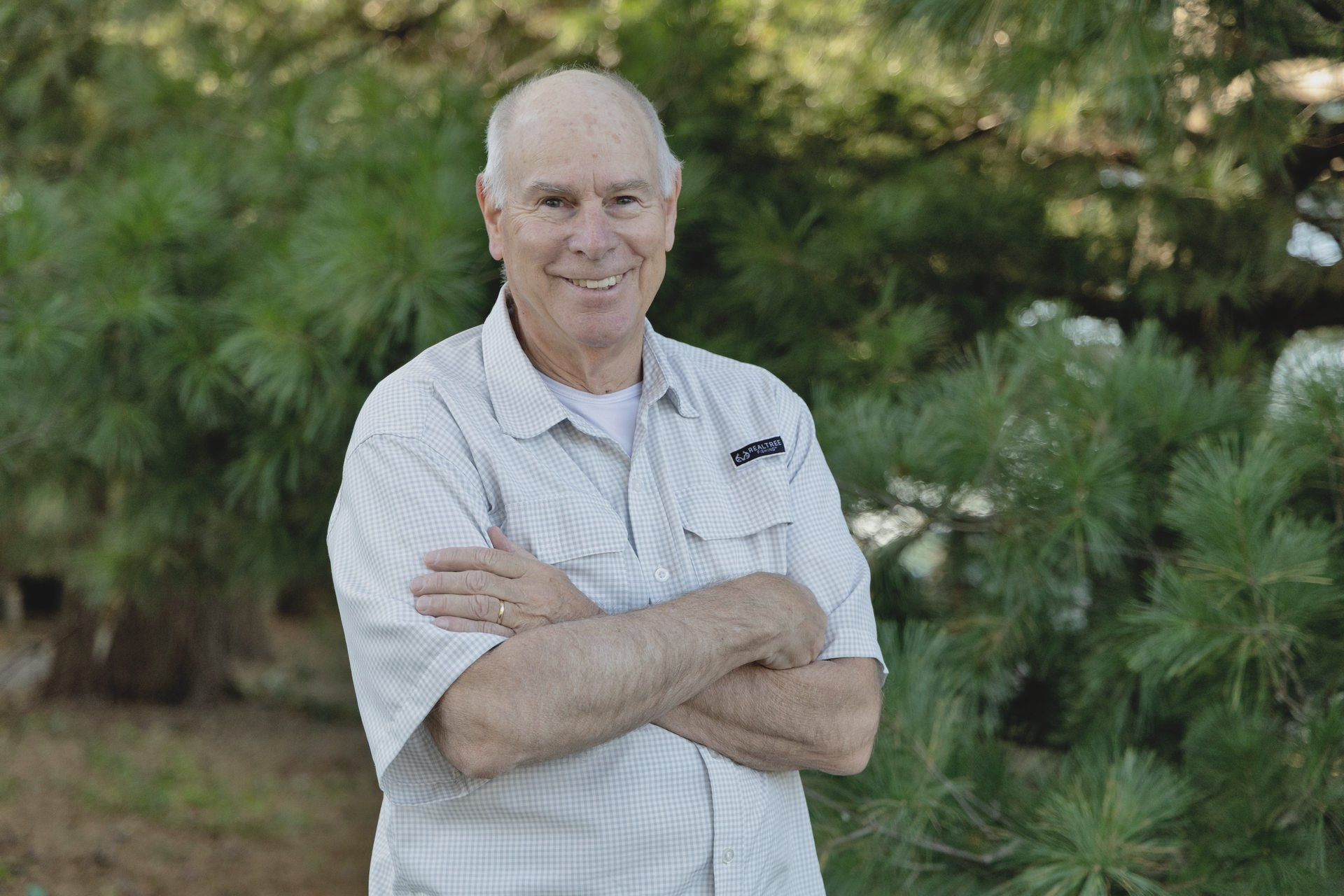 Man in light shirt with arms crossed, smiling, standing in front of green shrubbery.