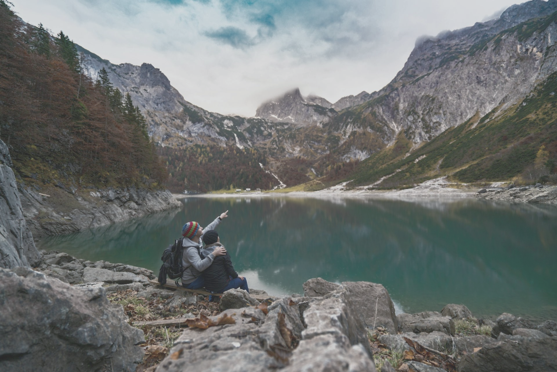 Couple sits by a lake, pointing towards mountains. Cloudy sky reflects in the water.