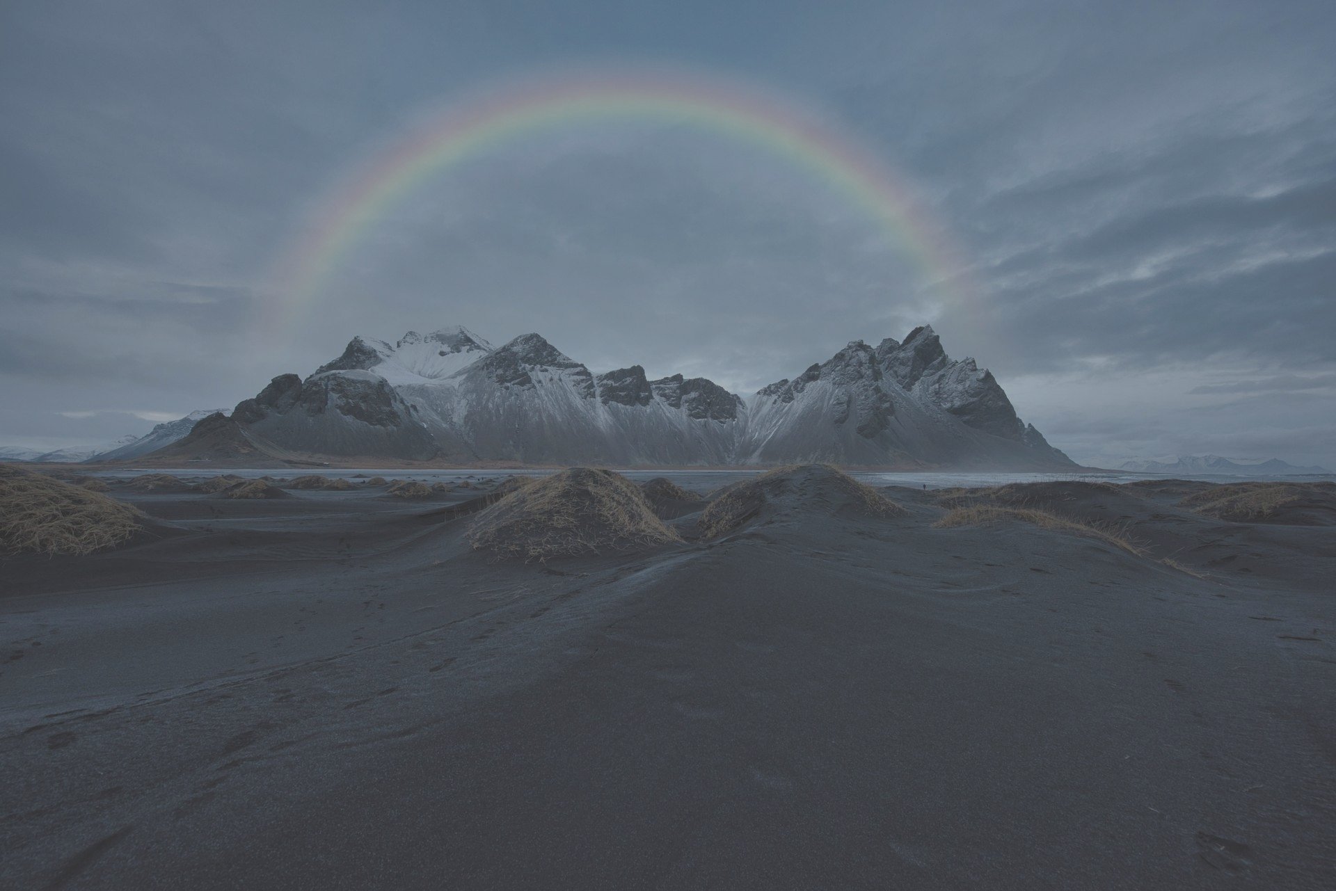 Rainbow arcs over snow-capped mountains and a dark, sandy foreground under a cloudy sky.