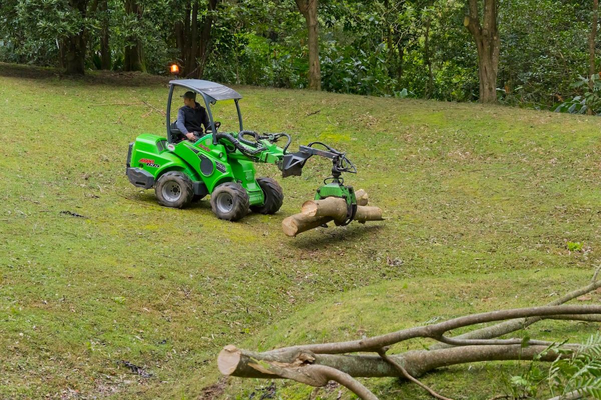 A Bulldozer Is Holding A Branch And Driving Down A Grass Field — Burnett Trees in Kiama, NSW