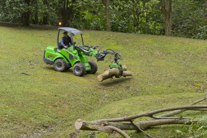 Green tractor with grapple arm lifting a log on a grassy hillside — Burnett Trees in Wollongong, NSW