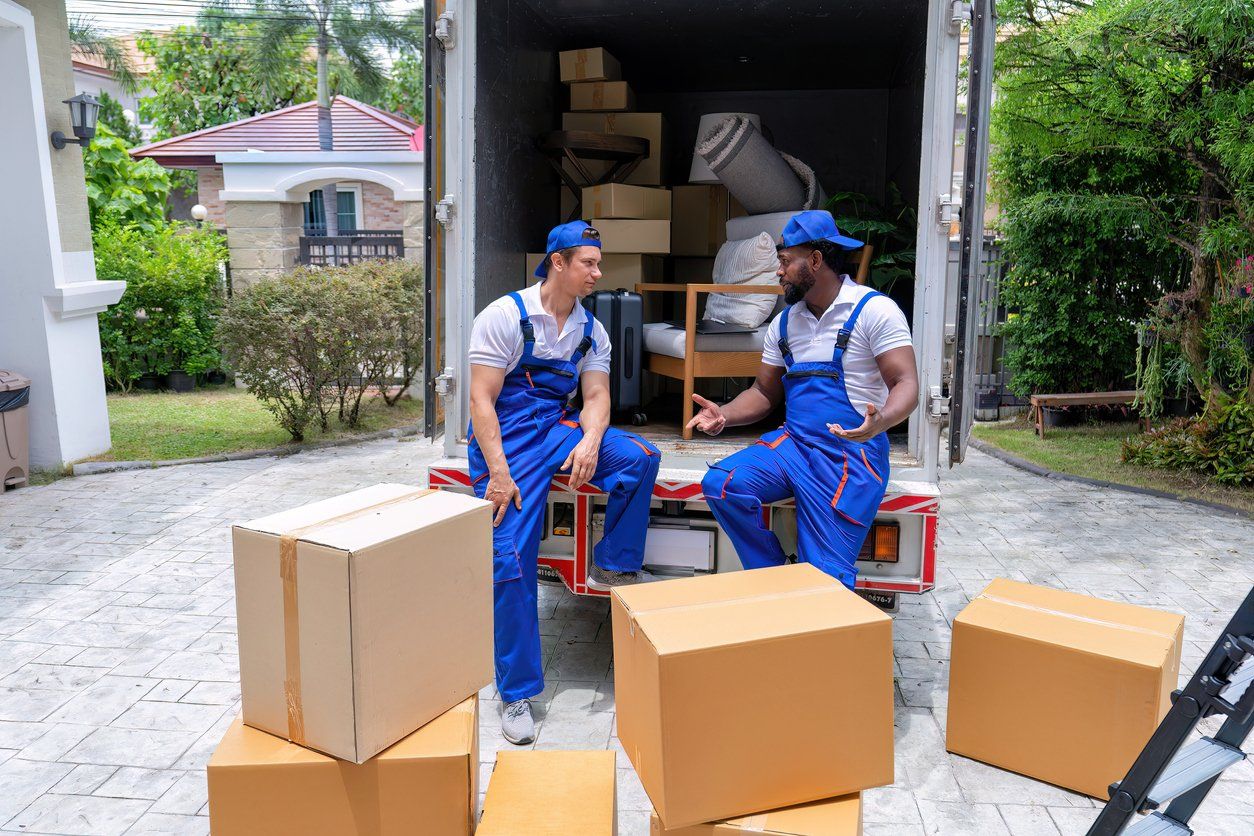 two movers in blue overalls sitting on the back of the moving truck with boxes on the driveway.