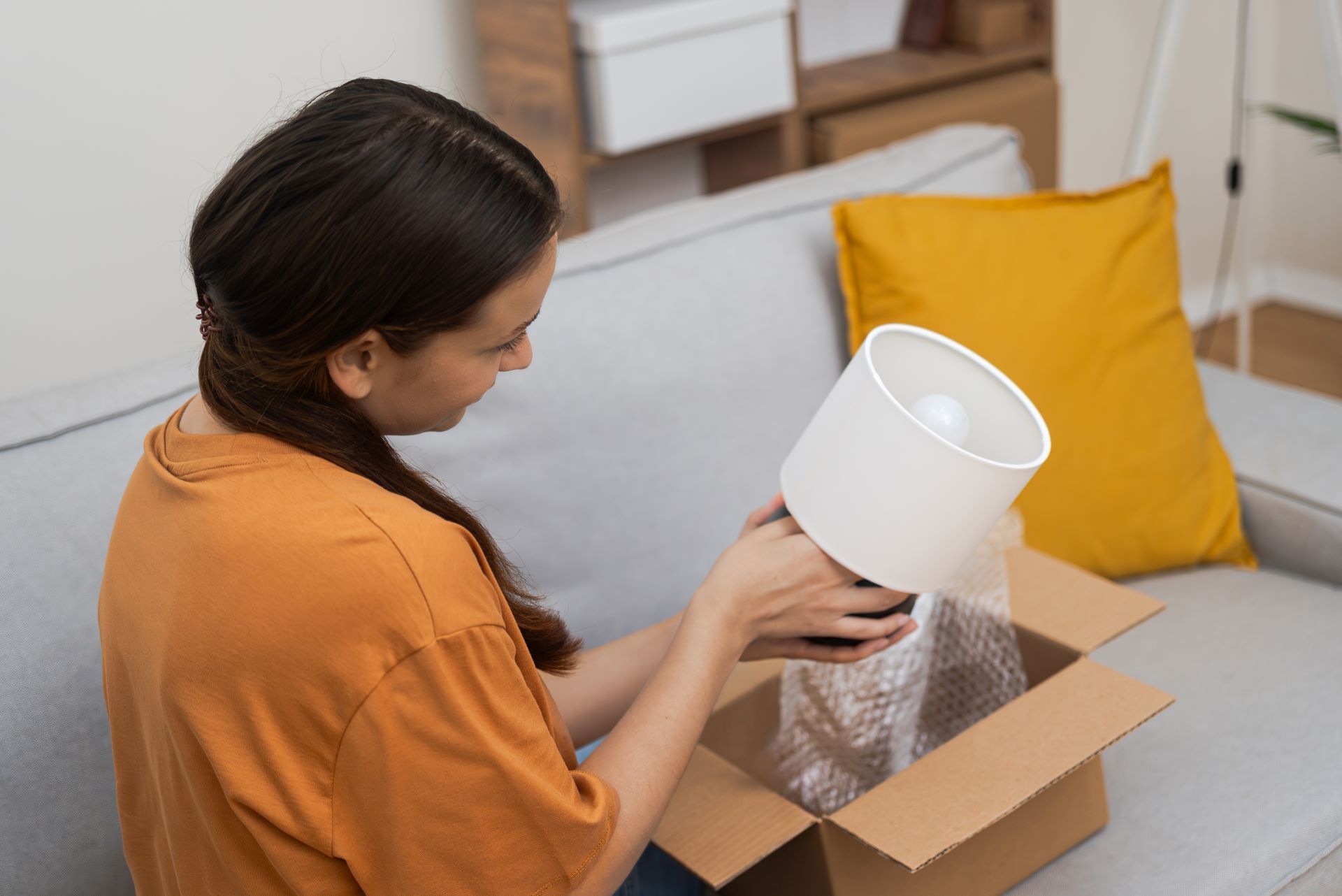 A woman is sitting on a couch holding a lamp shade in a cardboard box.