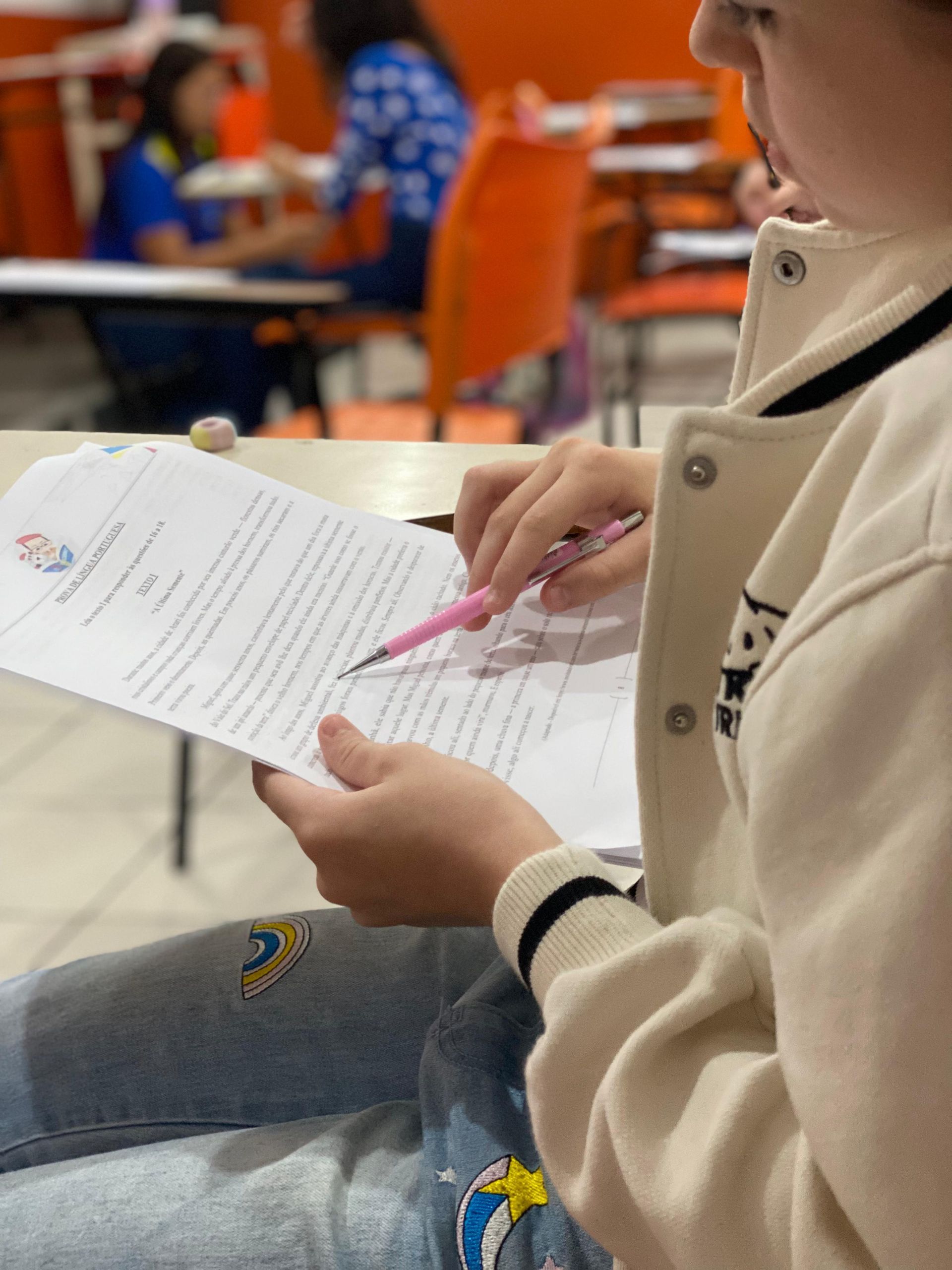Pessoa revisando um documento, segurando uma caneta rosa, sentada em um ambiente interno. Outras pessoas são visíveis ao fundo.