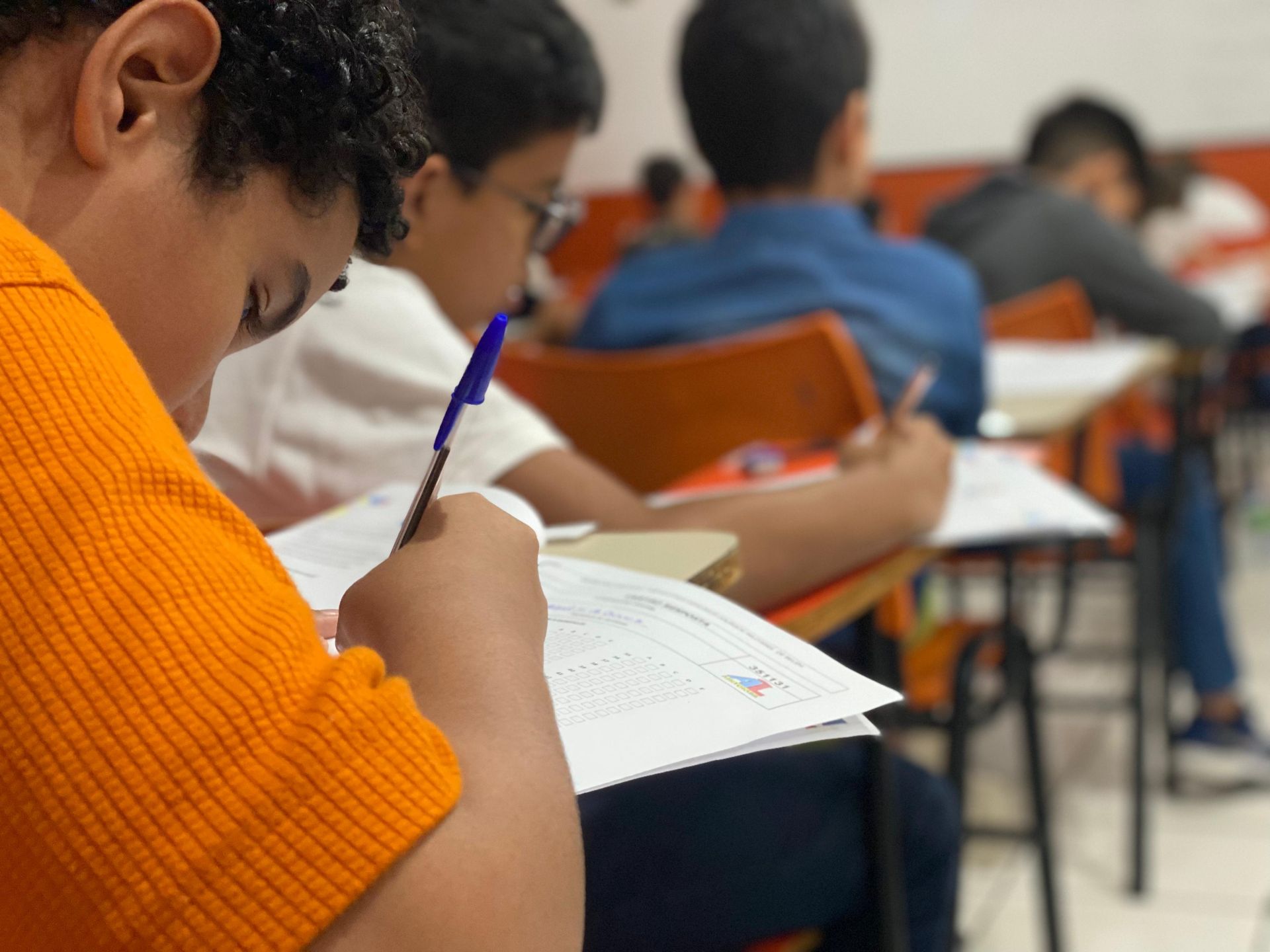 Alunos escrevendo em carteiras em uma sala de aula; um aluno de camisa laranja está em foco, segurando uma caneta.