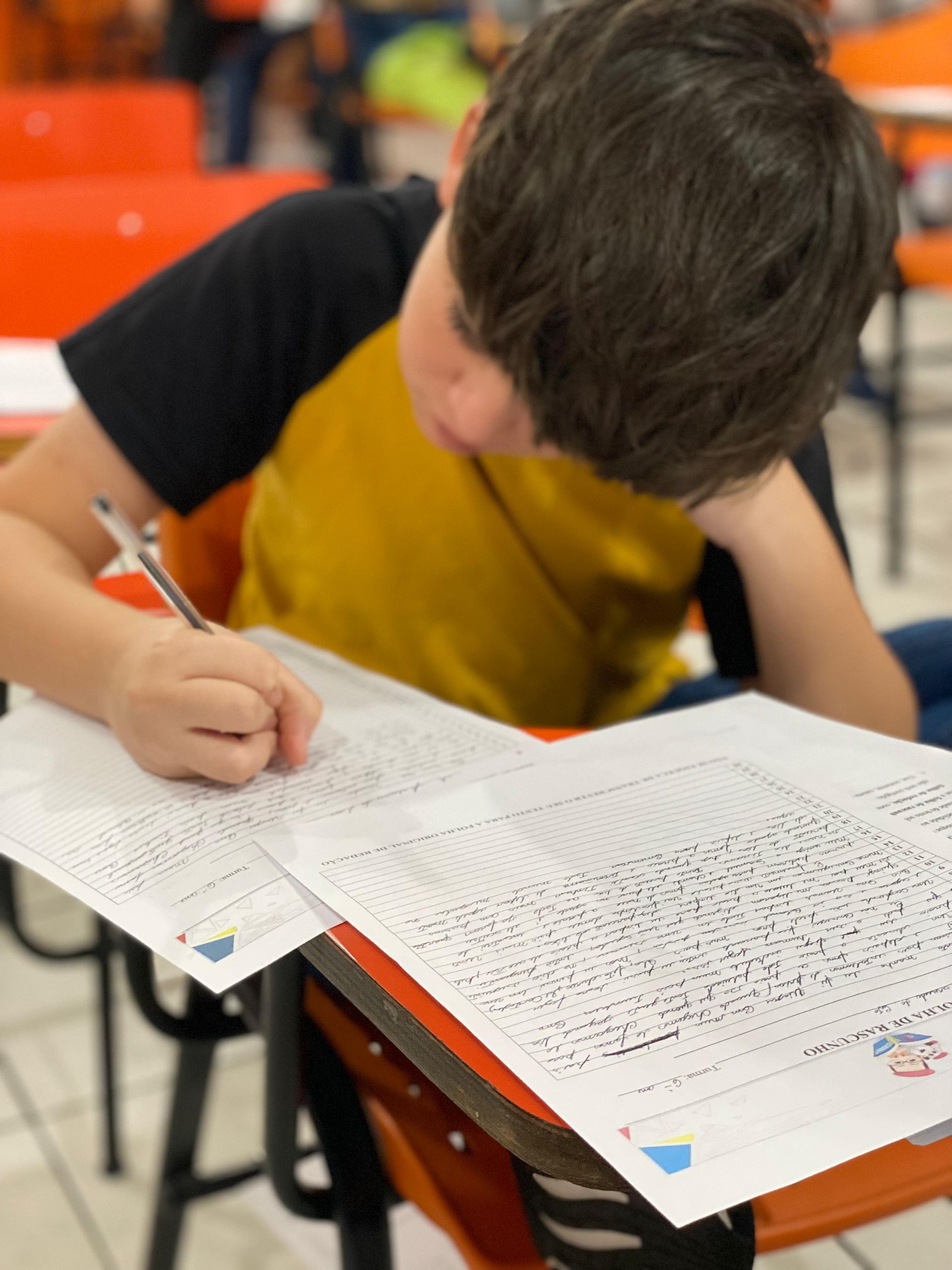Menino de cabelo escuro escrevendo em uma prova em sala de aula; ele está vestindo uma camisa amarela e inclinado para frente.