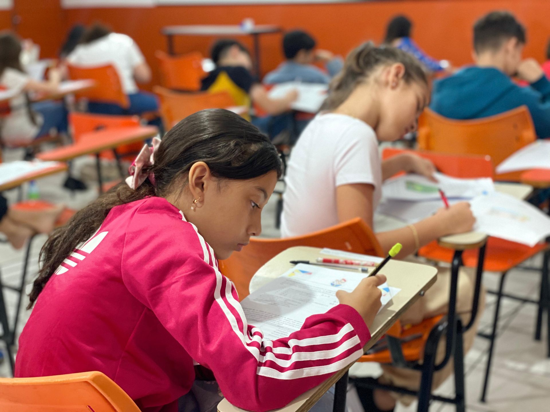Alunos fazendo uma prova em carteiras em uma sala de aula. Menina de jaqueta rosa escrevendo. Outros alunos ao fundo.