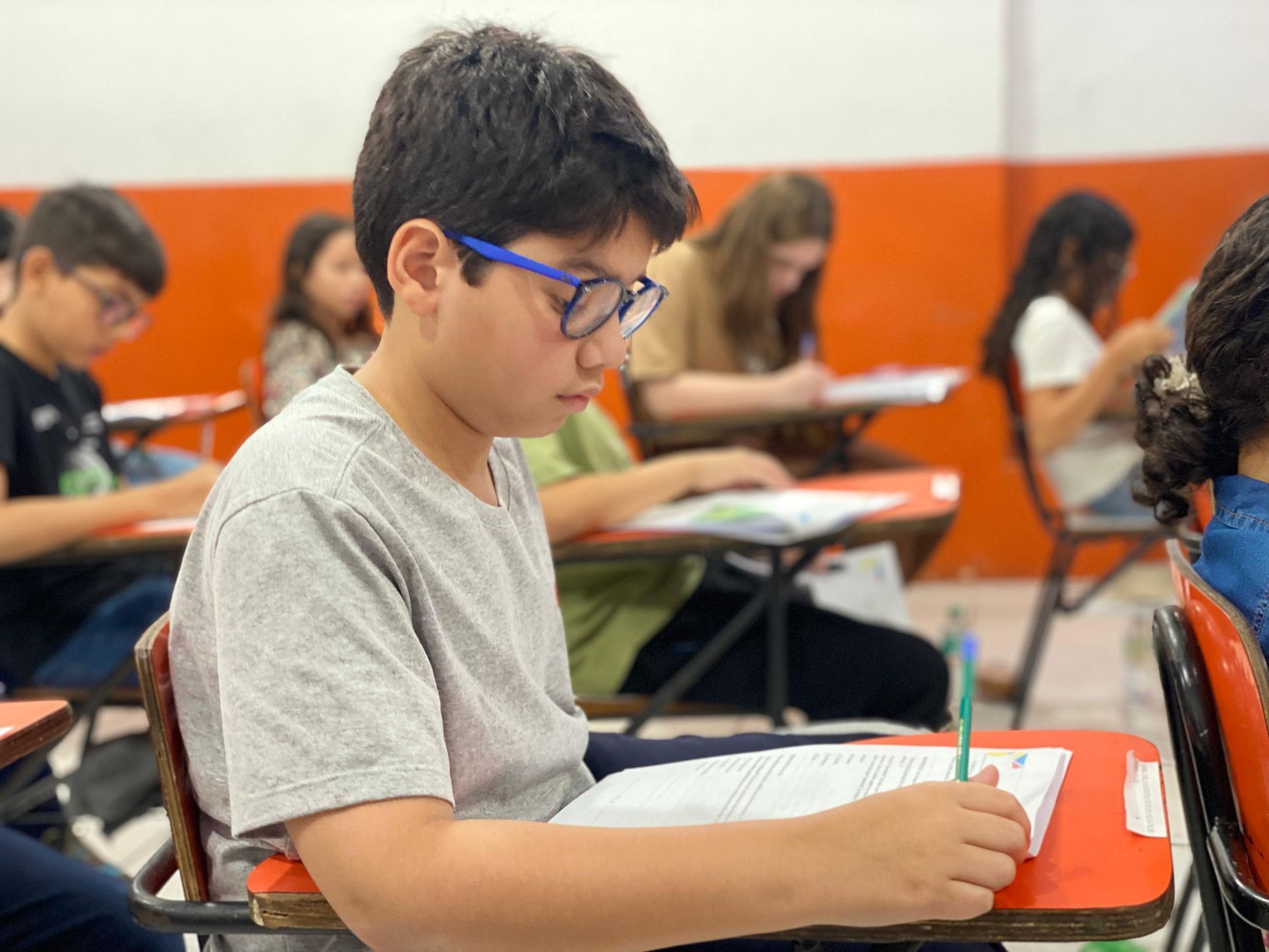 Menino de óculos escrevendo em uma mesa durante uma prova em uma sala de aula com outros alunos.