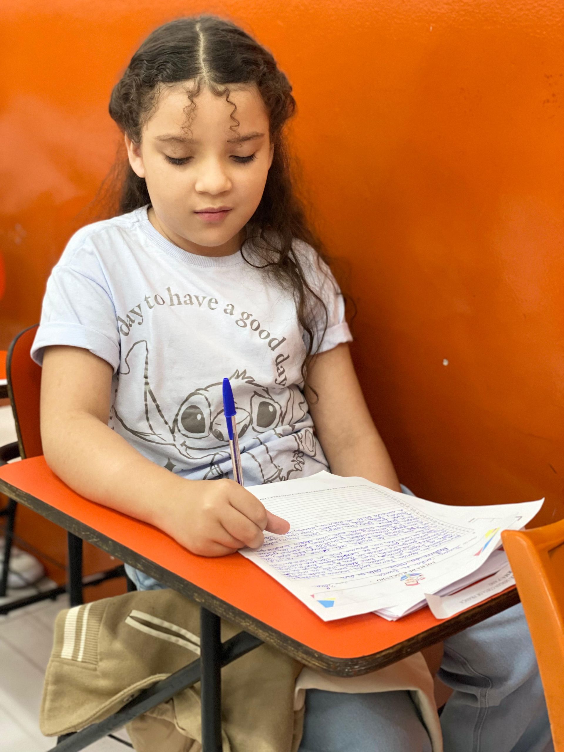 Menina de cabelos cacheados escrevendo em uma mesa, olhando para baixo. Parede e mesa laranja, vestindo uma camisa azul-clara.