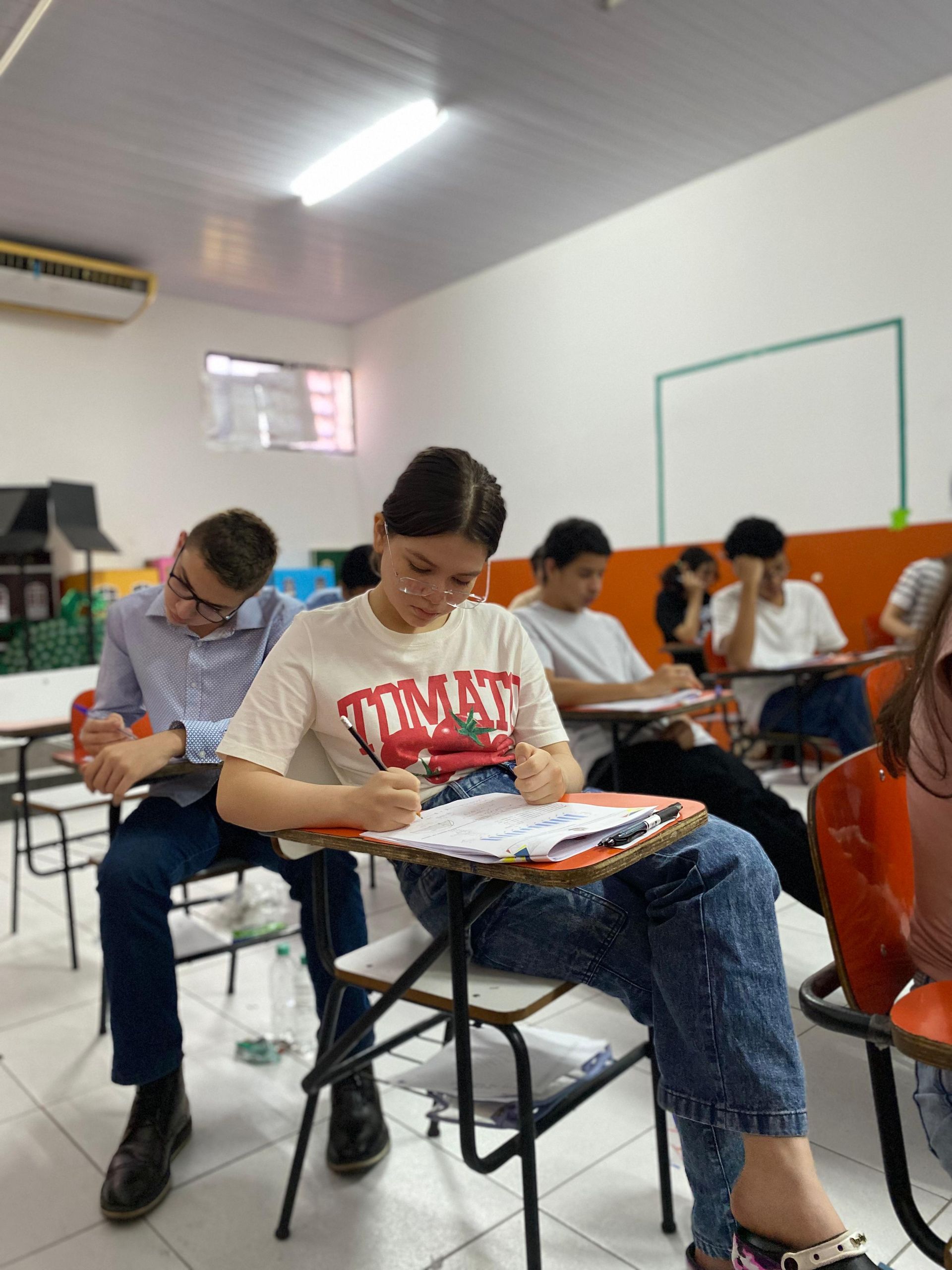 Alunos fazendo uma prova em uma sala de aula. Menina no centro escrevendo, outros sentados em carteiras, concentrados.
