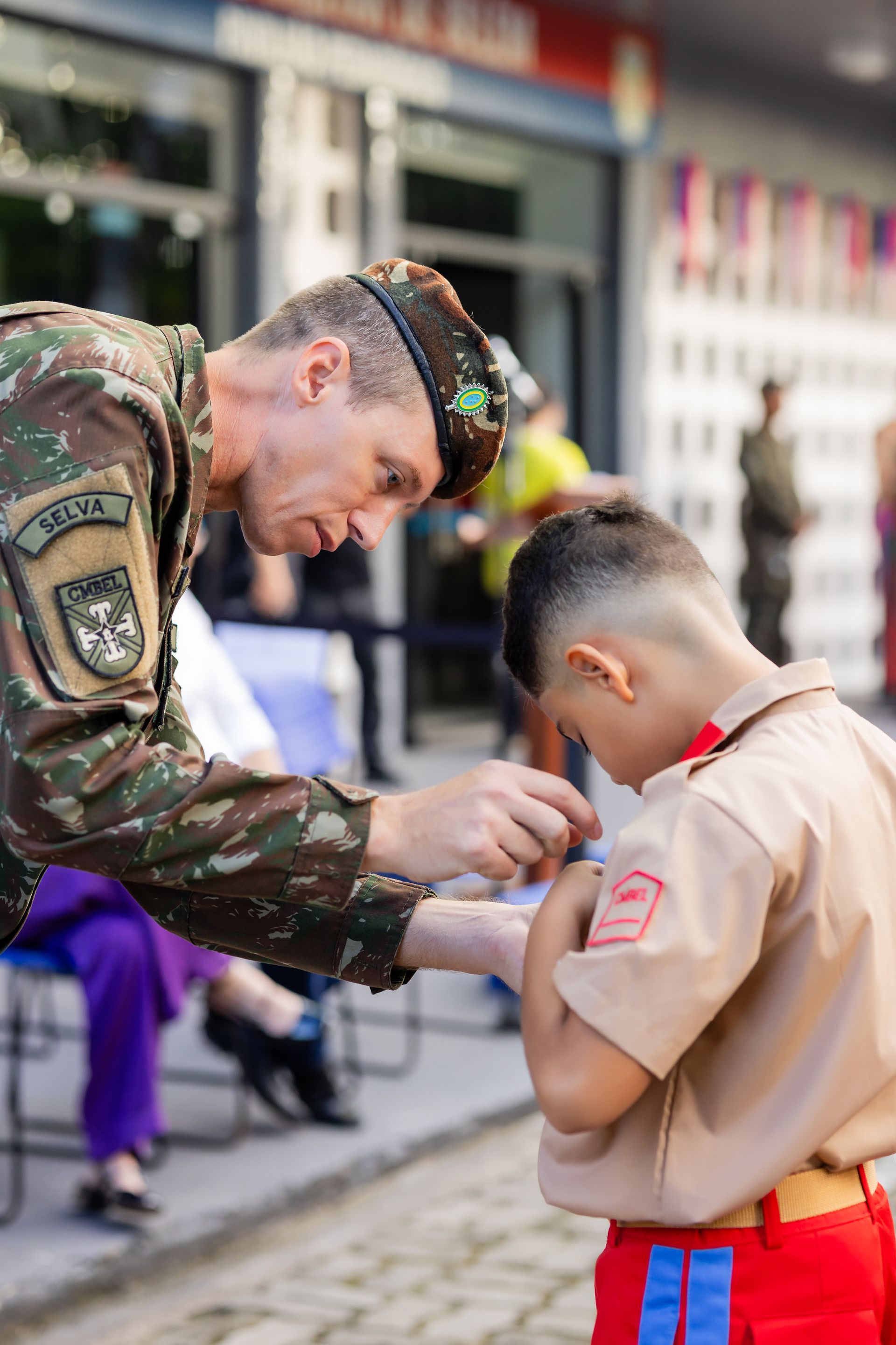 Soldado prendendo um emblema no braço de um garoto de uniforme bege, ao ar livre.