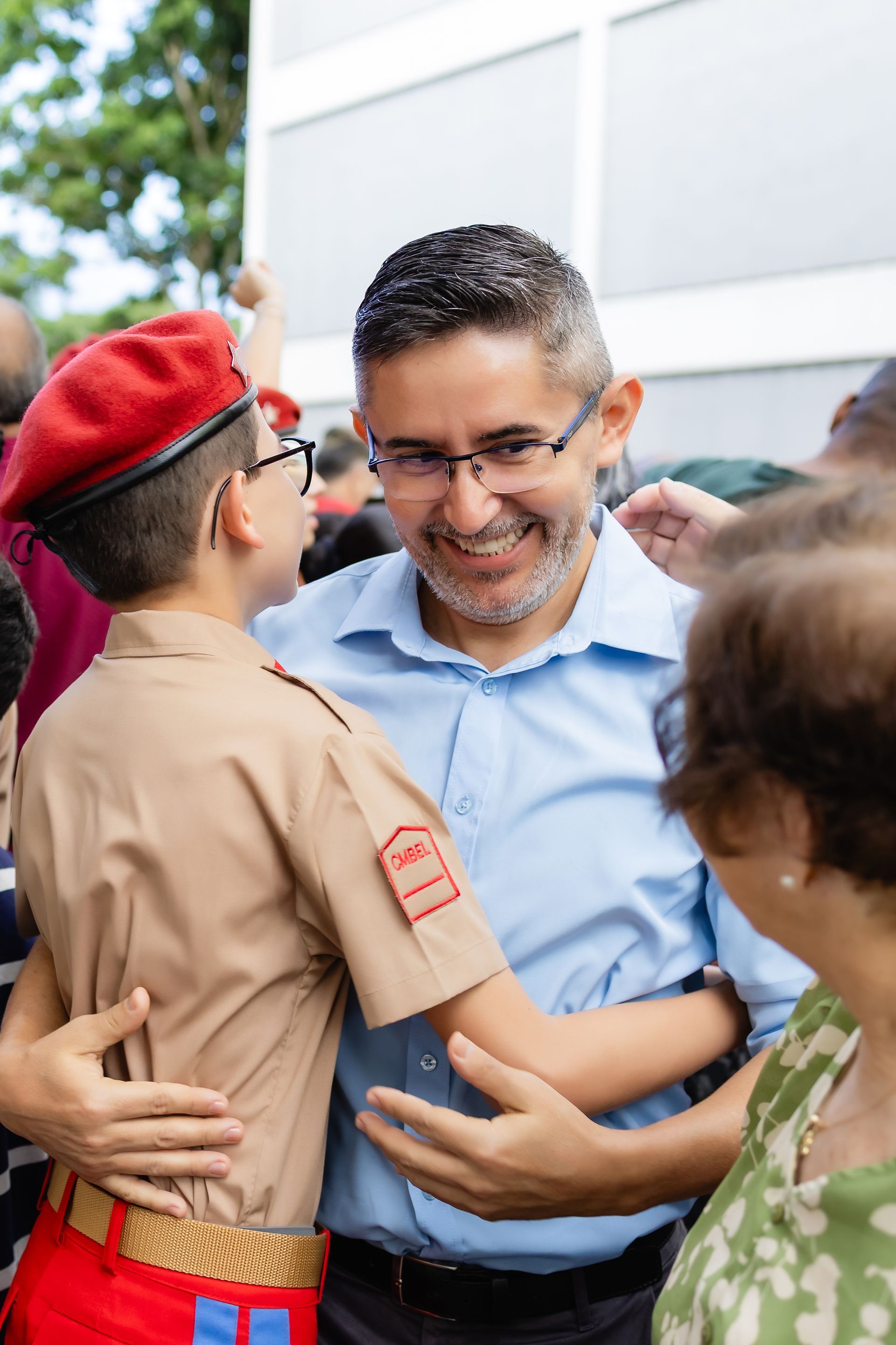 Homem abraçando um menino de boina vermelha e uniforme. Sorrindo, possivelmente ao ar livre.